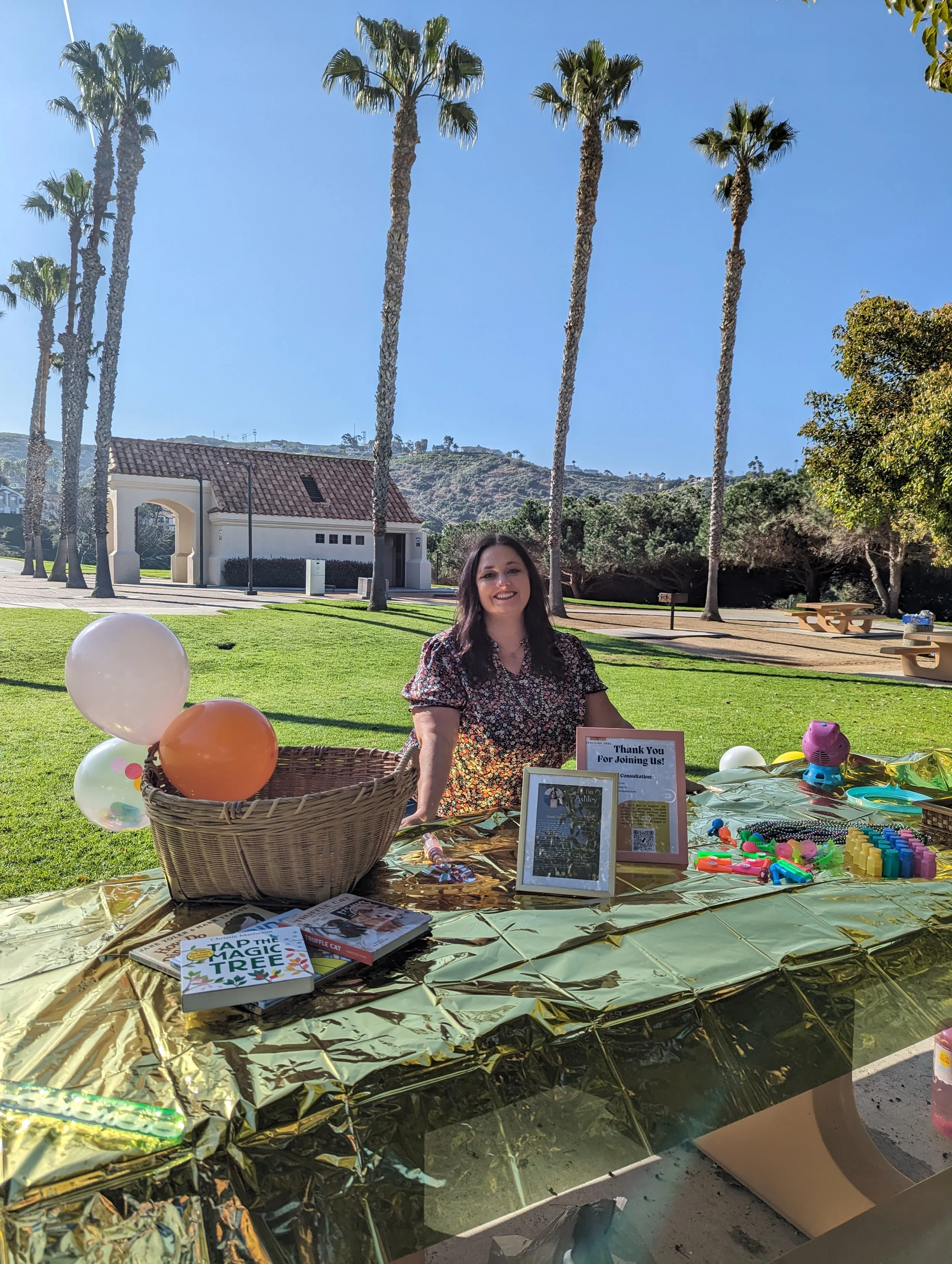 A woman is sitting at an outdoor table decorated with balloons and toys, smiling. The table has books, signs, and party favors. There are palm trees, a building, and hills in the background under a clear blue sky.