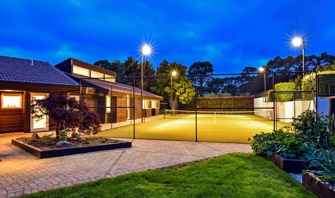 Modern home exterior with tennis court and floodlights behind fence.