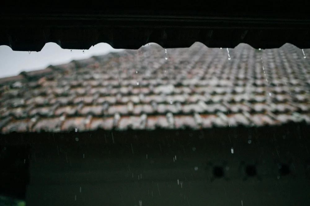A close-up, shallow-depth-of-field shot of raindrops dripping from the edge of a dark roof eaves with a blurred weathered tile roof in the background.