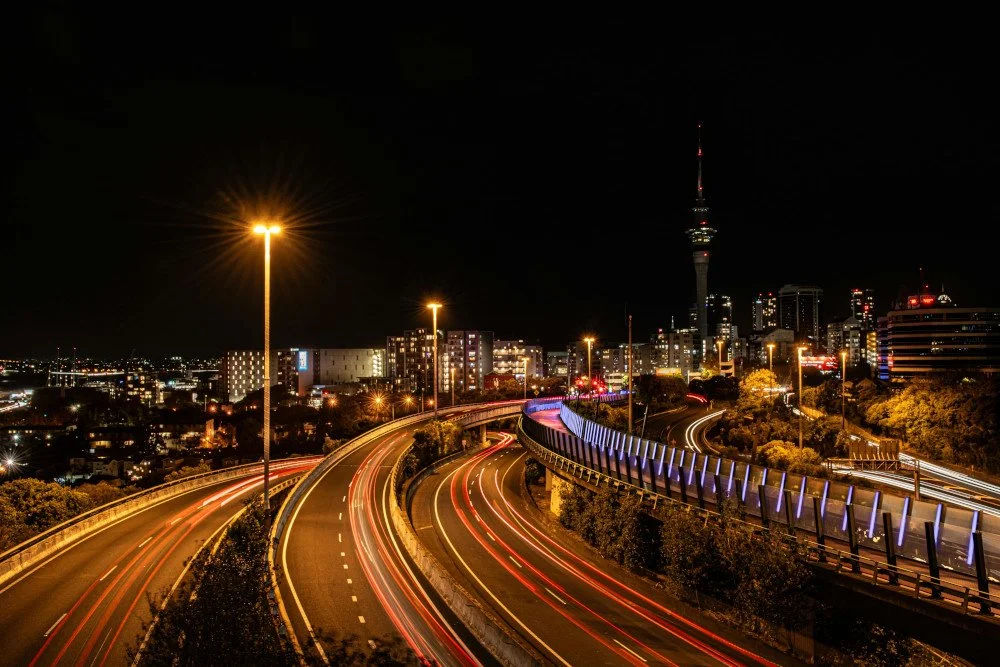 A long-exposure night photograph of the Auckland skyline featuring the Sky Tower and light trails from traffic on a curving motorway.