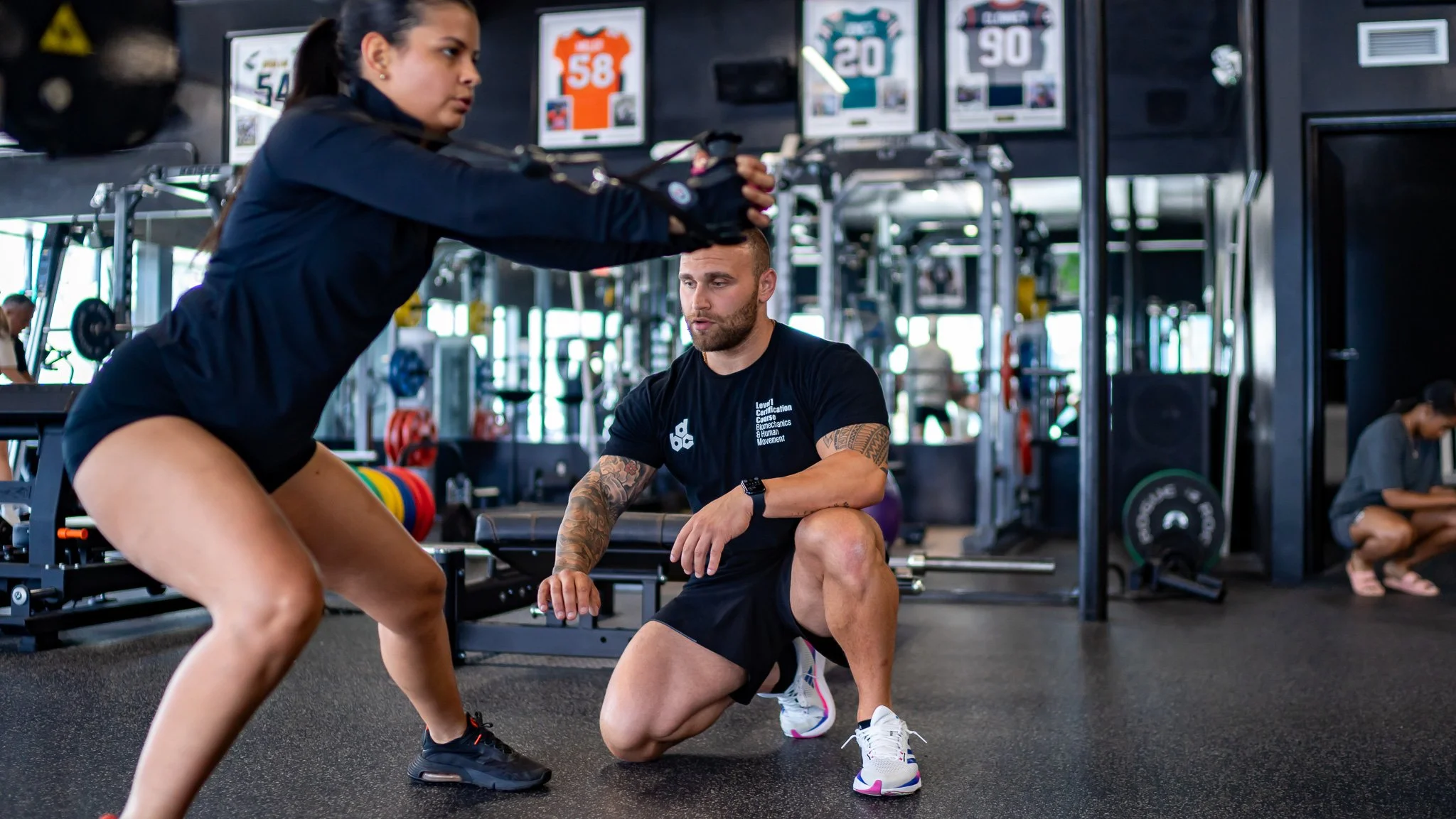 A woman with dark hair in a ponytail wearing a black long-sleeve shirt and shorts is doing a squat exercise with a weight overhead, assisted by a male trainer with a beard and tattoos, kneeling on one knee in a gym.