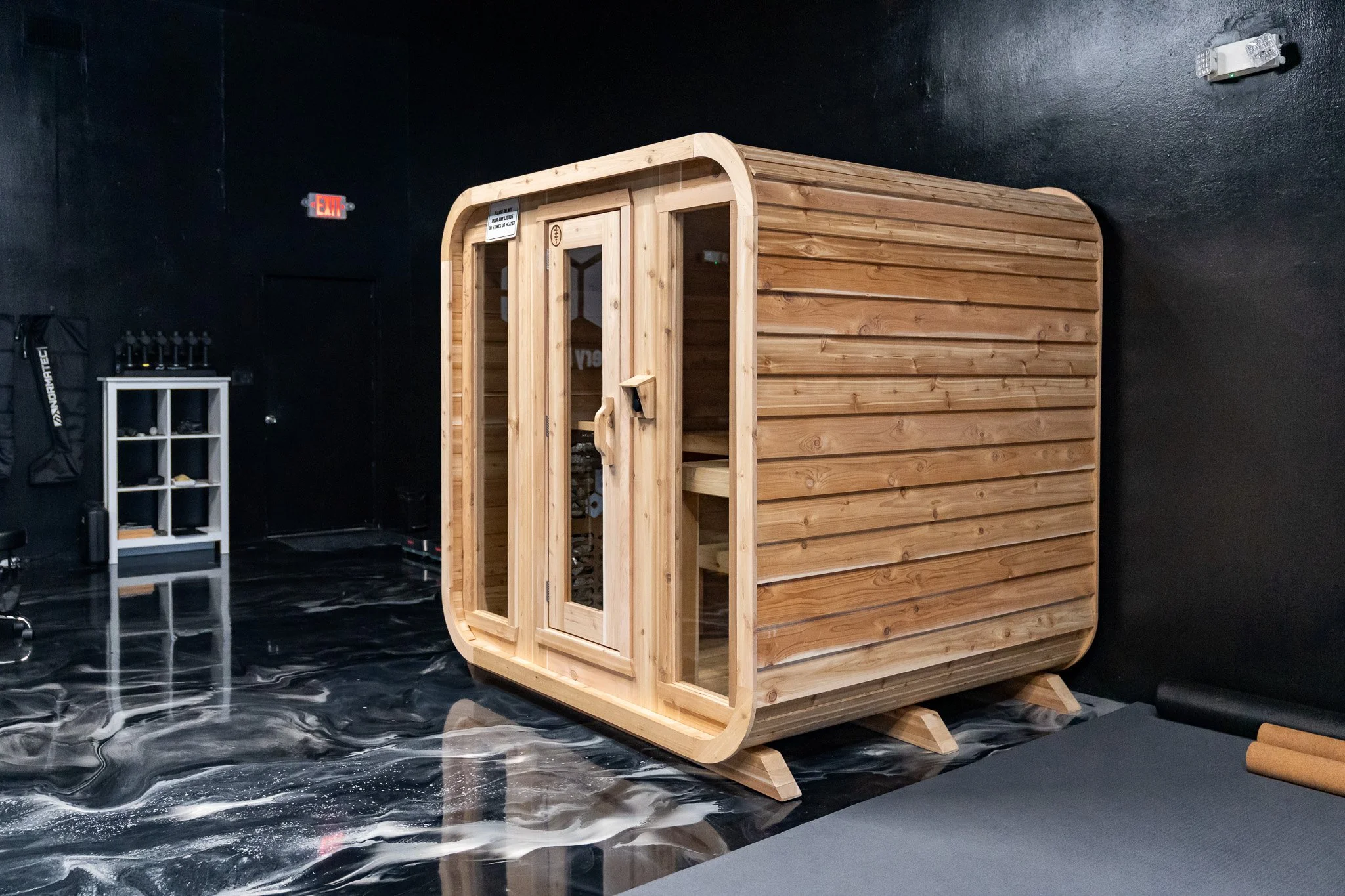 Indoor sauna with wooden exterior, glass door, situated on a black marble floor surrounded by fitness and wellness equipment.