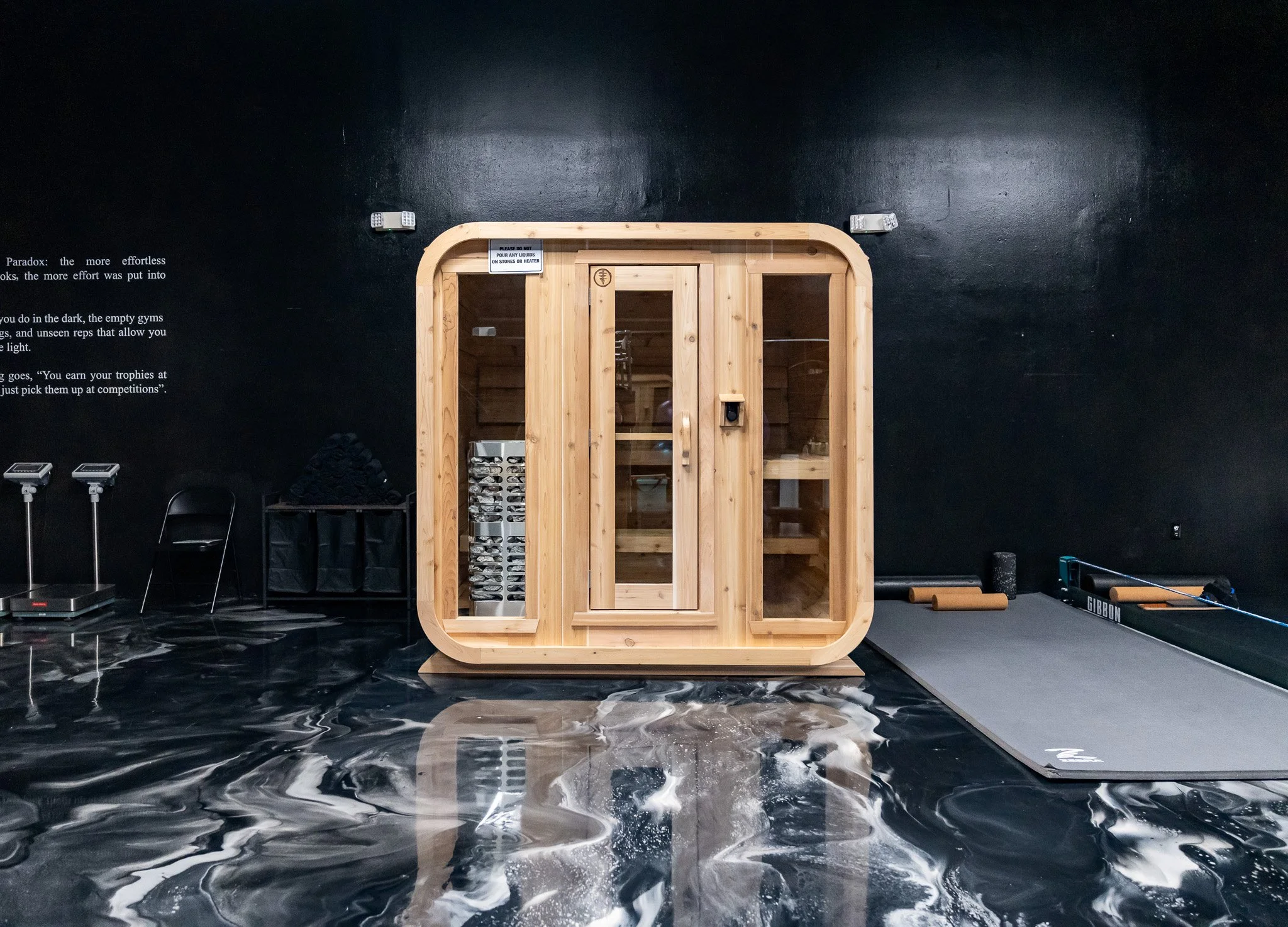 Wood sauna with glass door in fitness studio with black marbled flooring and black walls.