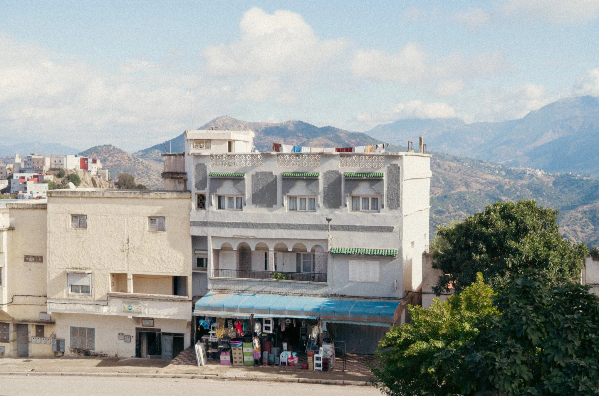 A white multi-story building with small windows and green awnings, a storefront with a blue awning, and laundry hanging on the rooftop, set against a mountainous landscape with other buildings and a large green tree in the foreground.