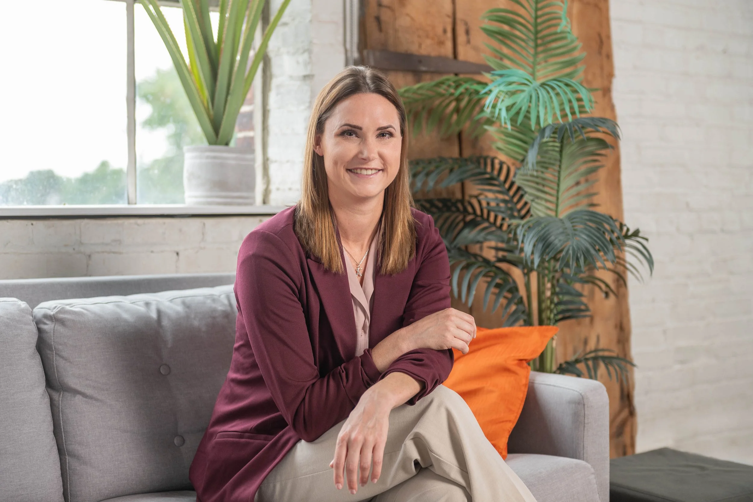 A woman with shoulder-length brown hair smiling, sitting on a gray couch in a modern, industrial-style living room, with a large green plant and an orange pillow behind her, and a window with natural light.