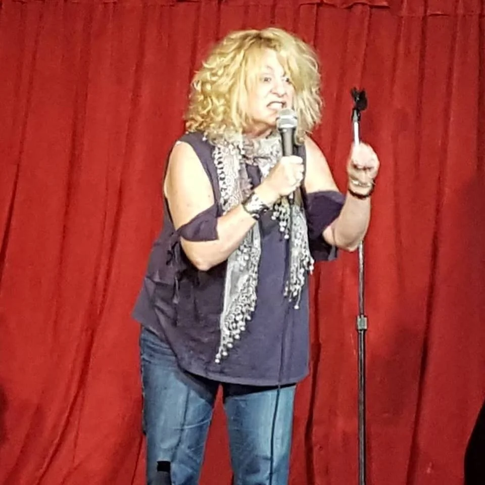 Woman performing stand-up comedy on stage with red curtains, holding a microphone, wearing a scarf and casual clothes.