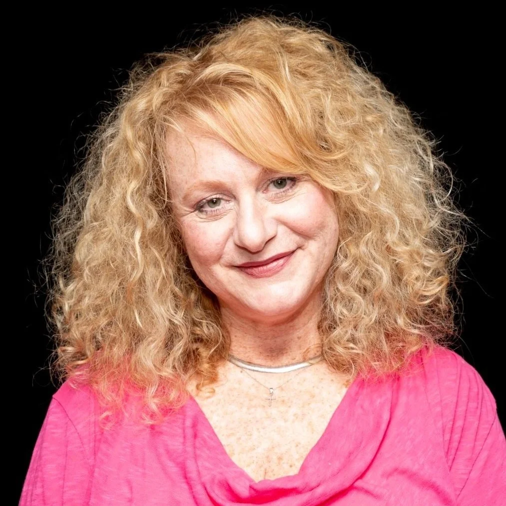 Person with curly blonde hair, wearing a pink top, smiling against a black background.