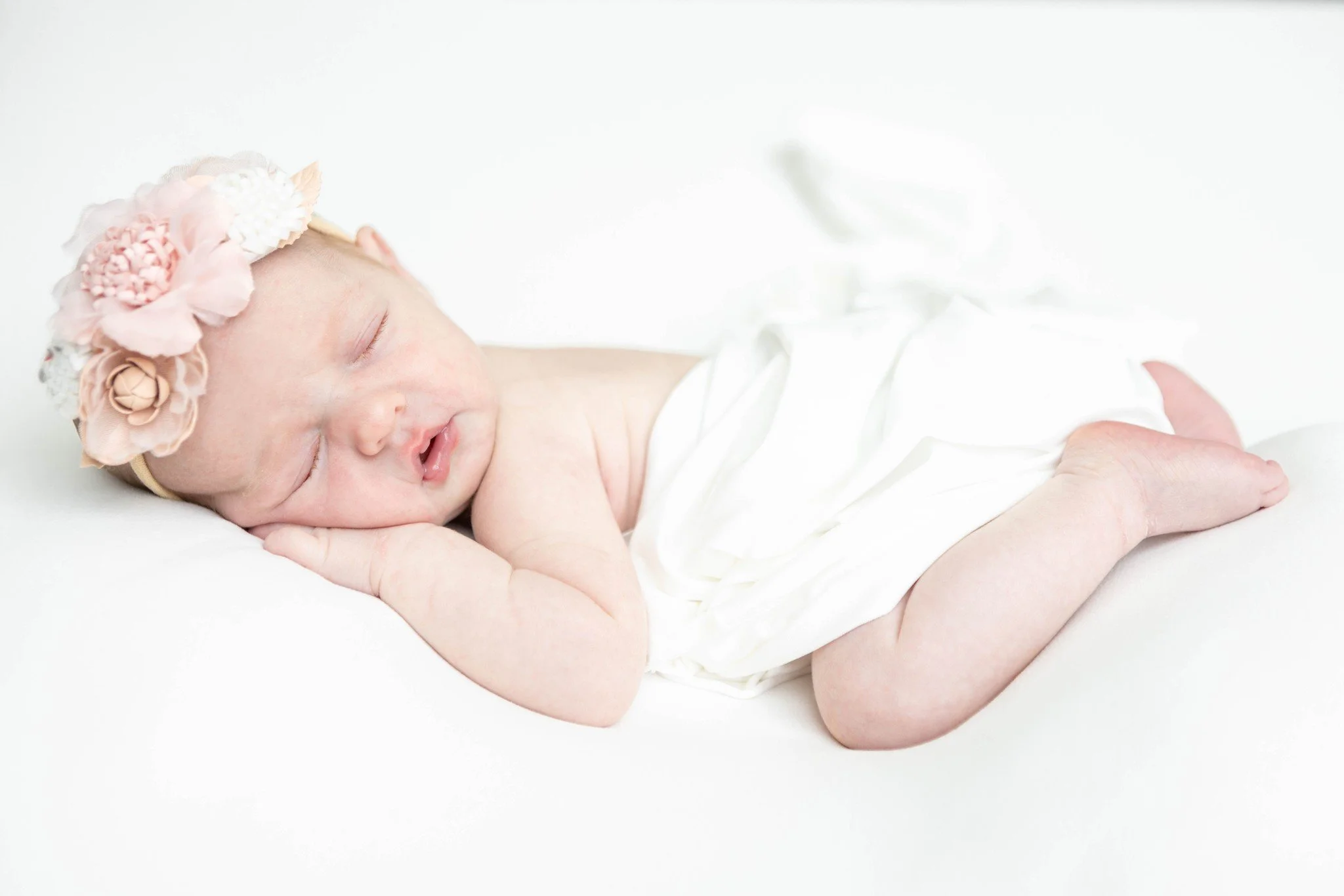 A sleeping newborn baby wearing a floral headband, lying on a white surface, wrapped in a white cloth.