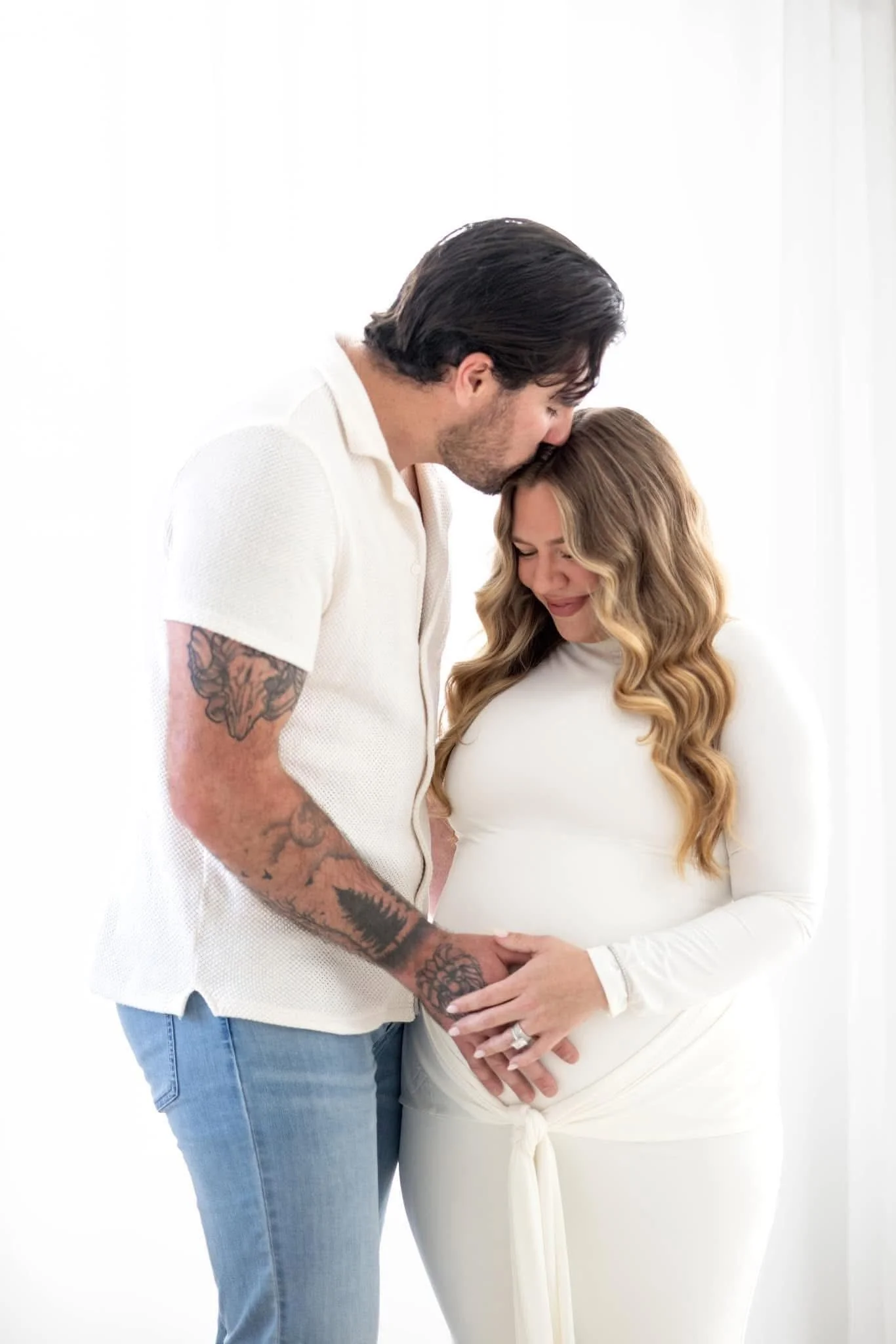 A pregnant woman with long blonde wavy hair in a white dress, with a man with dark hair and tattoos on his arm, kissing her on the forehead and holding her belly.
