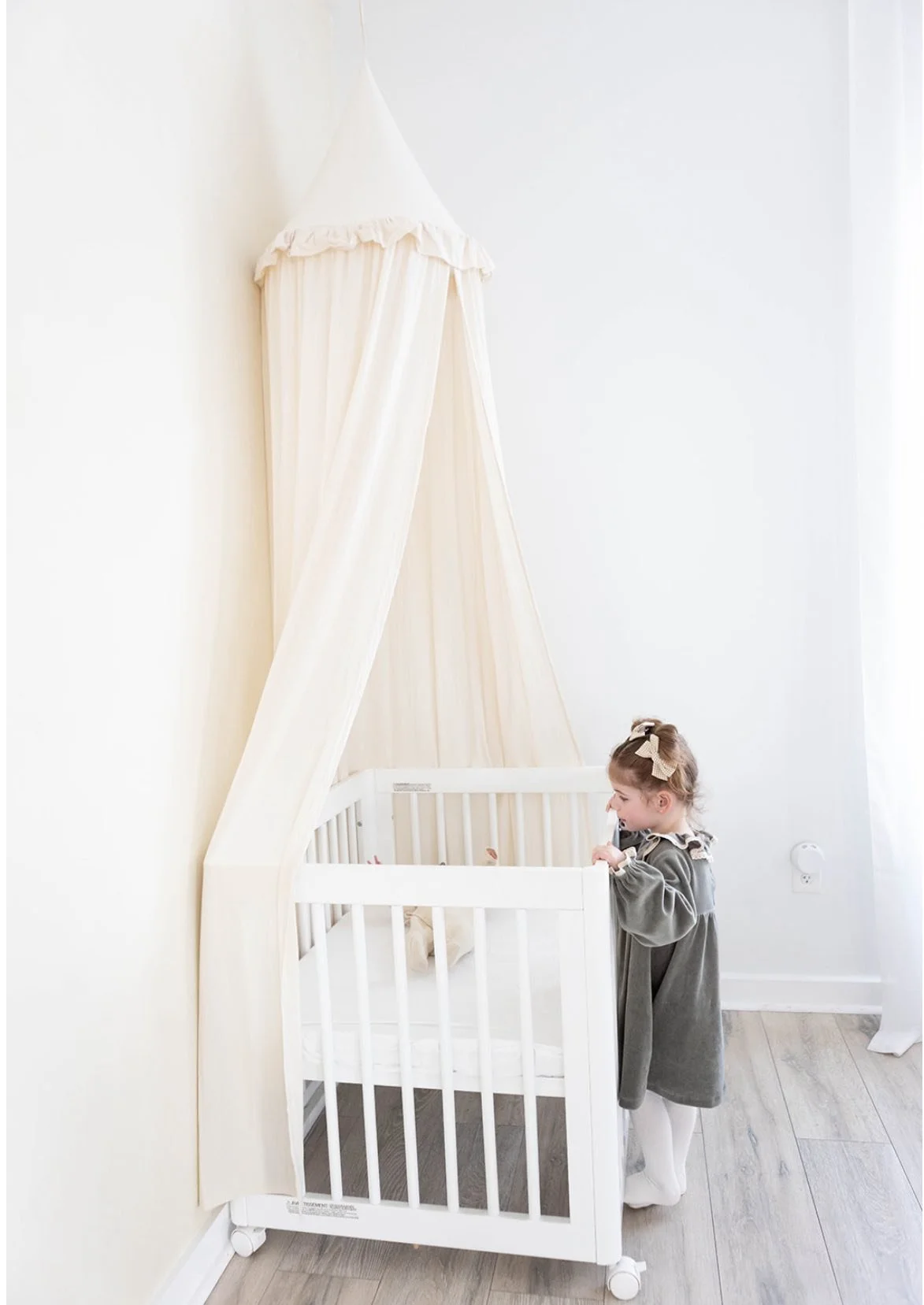 A young girl with a bow in her hair standing next to a white crib with a canopy in a bright, minimalistic room with light-colored wood flooring.