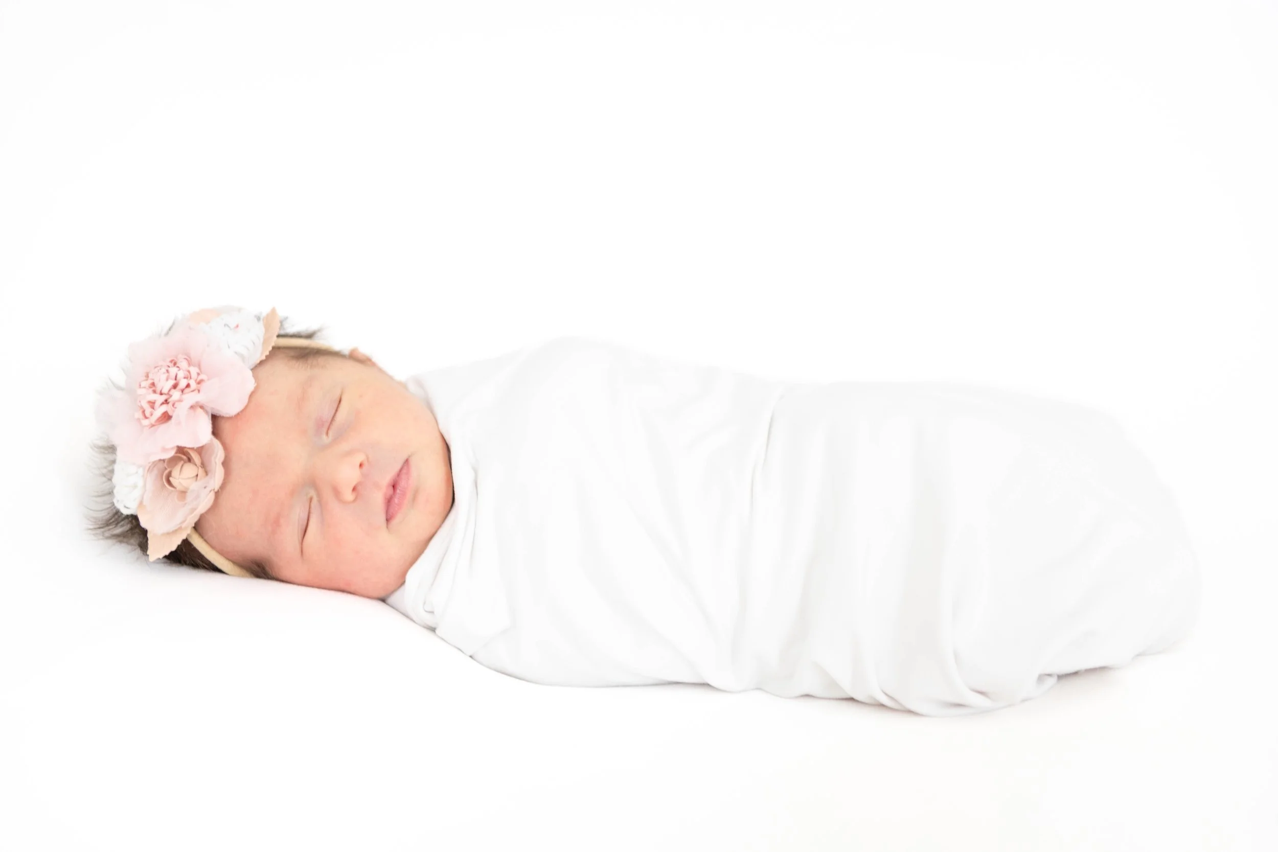 A sleeping baby girl wearing a floral headband, lying on a white surface wrapped in a white blanket.