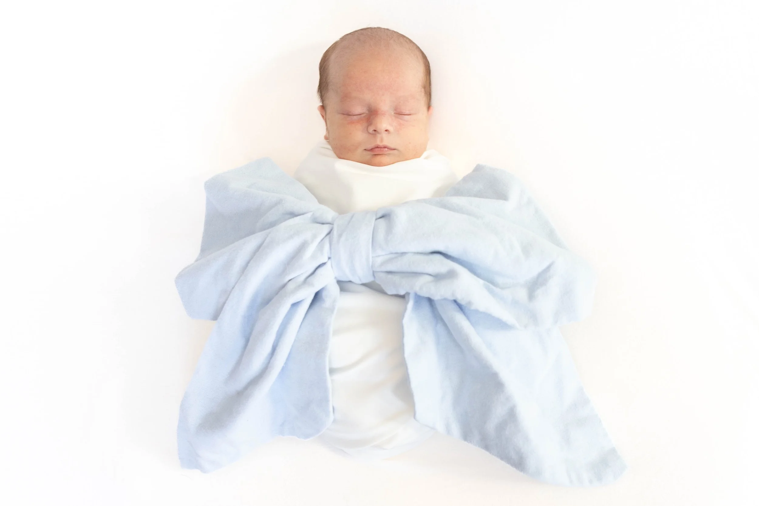 Newborn baby with closed eyes wrapped in white cloth and blue blanket, lying on white background.