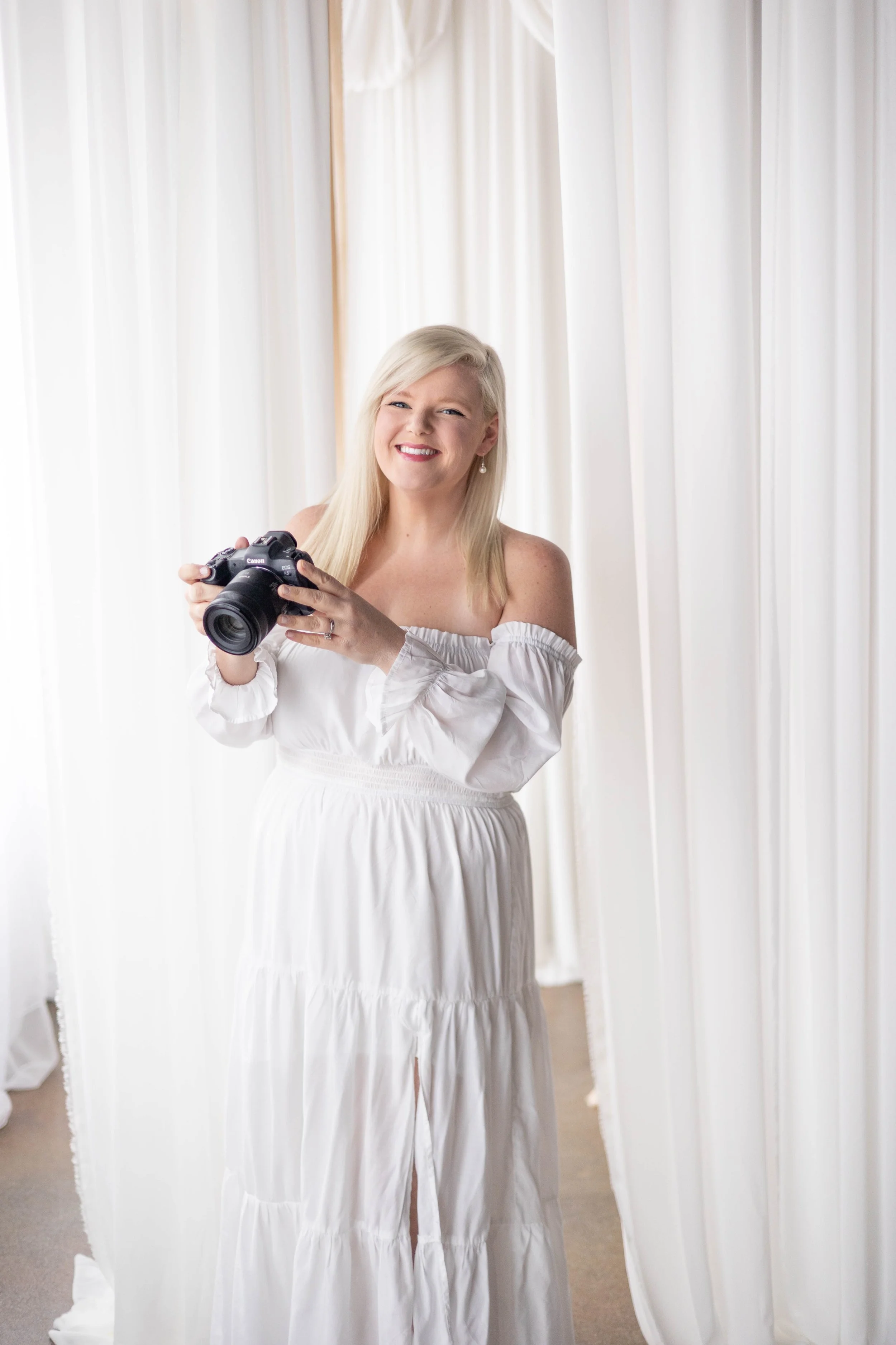A young woman in a white off-the-shoulder dress smiling and holding a camera in front of white curtains.