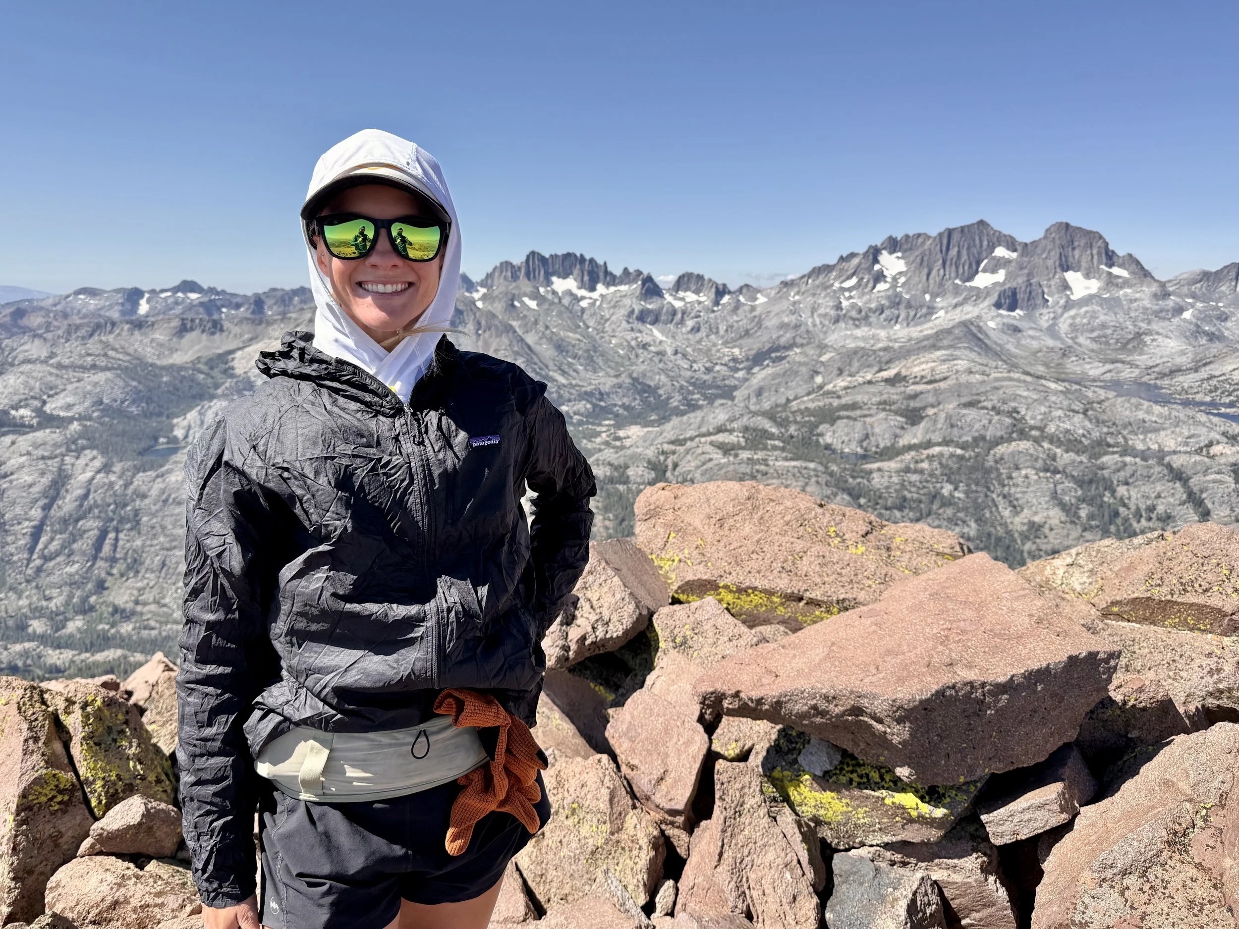 A smiling woman in outdoor mountain gear stands on rocky terrain with snow-capped mountains in the background under a clear blue sky.