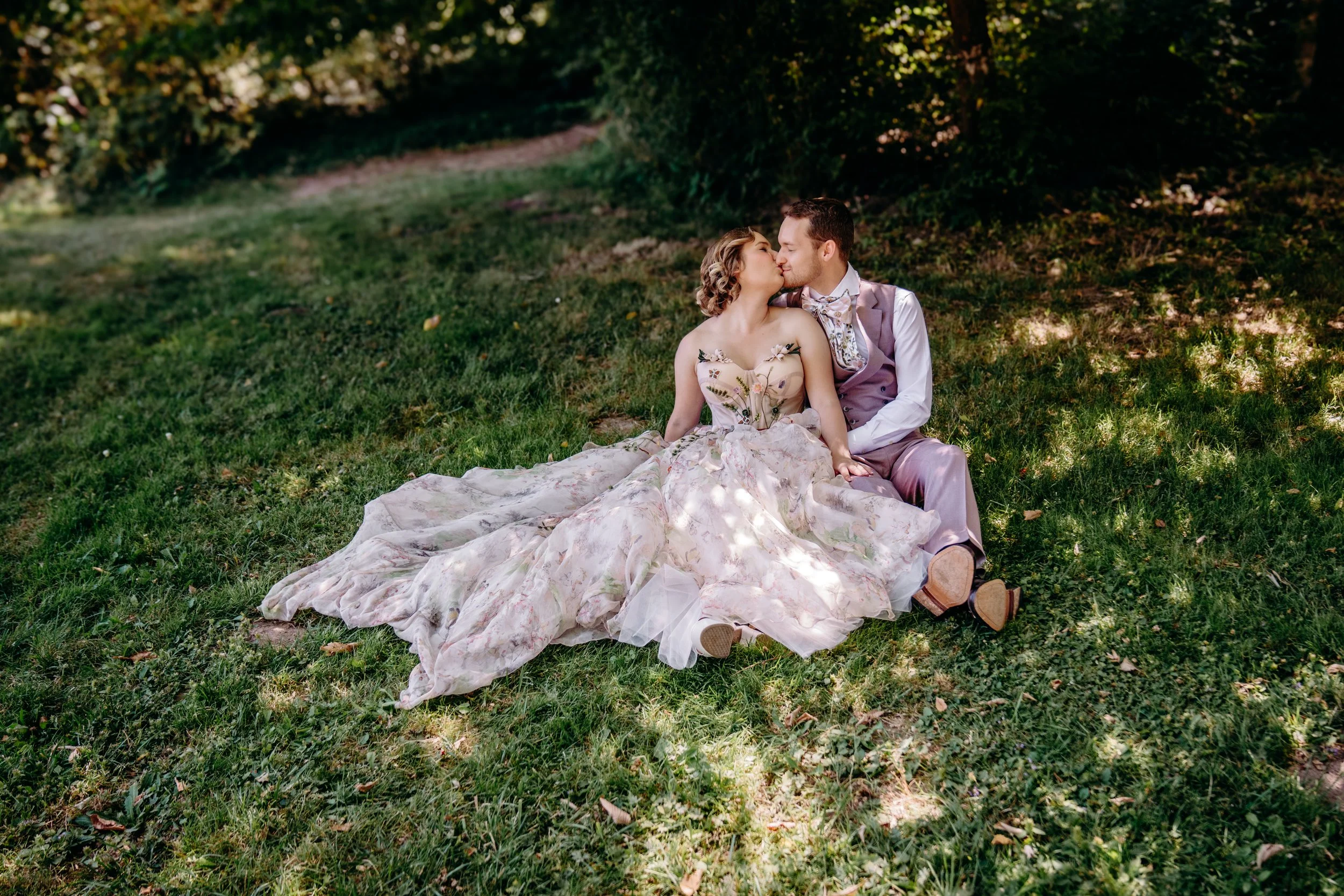 A couple dressed in vintage wedding attire sitting on grass, sharing a kiss outdoors under trees.