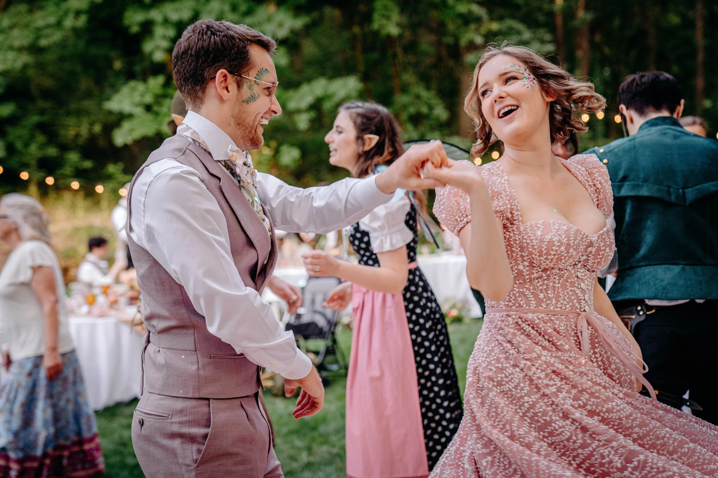 People dancing and having fun outdoors at a party or wedding reception, surrounded by trees and string lights.