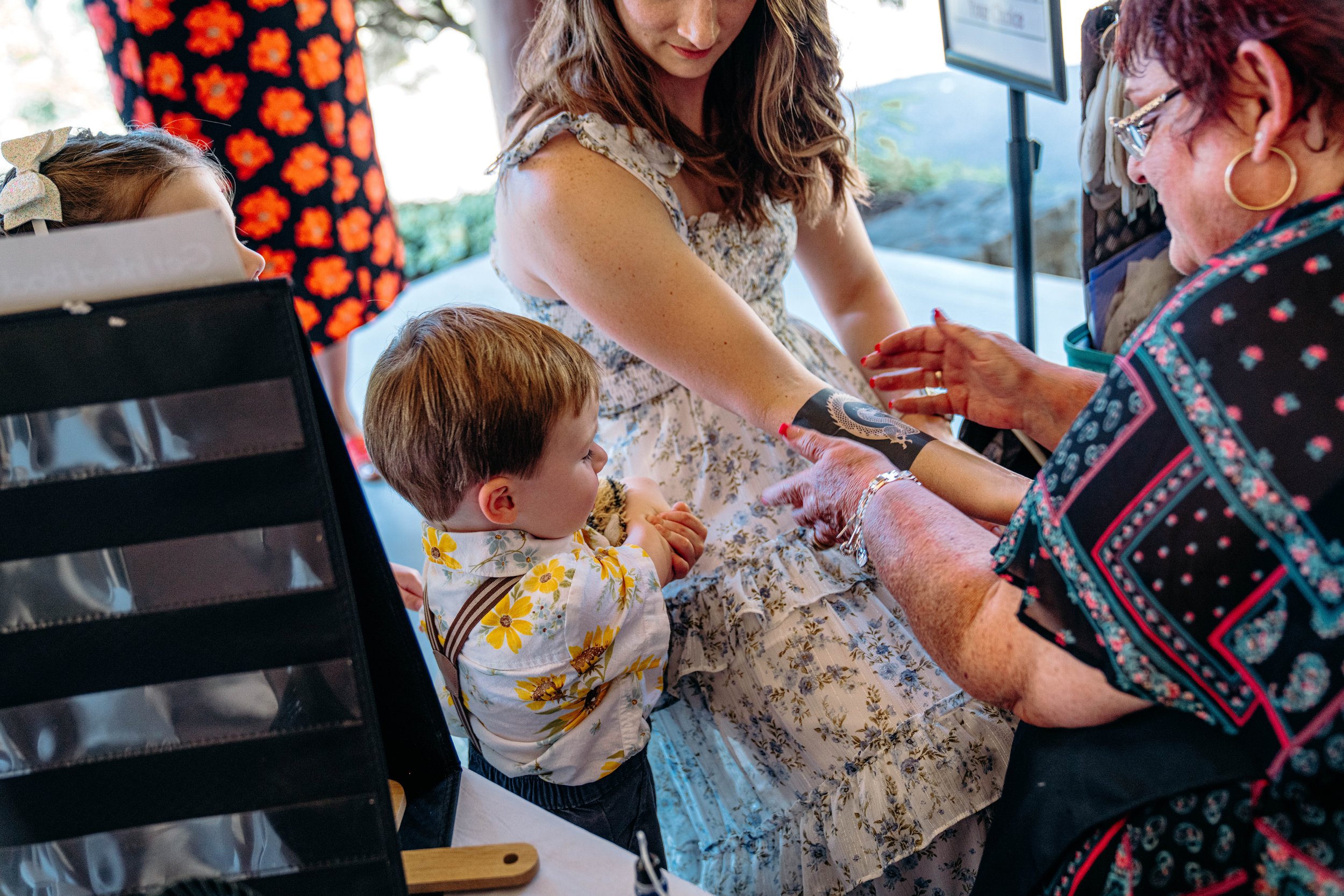 A young boy in a floral shirt is standing in a line, watching a woman in glasses and a floral blouse place a tattoo on a girl’s arm. The girl, in a floral dress, looks down while another woman, in a black and pink blouse, prepares to do the tattoo. M