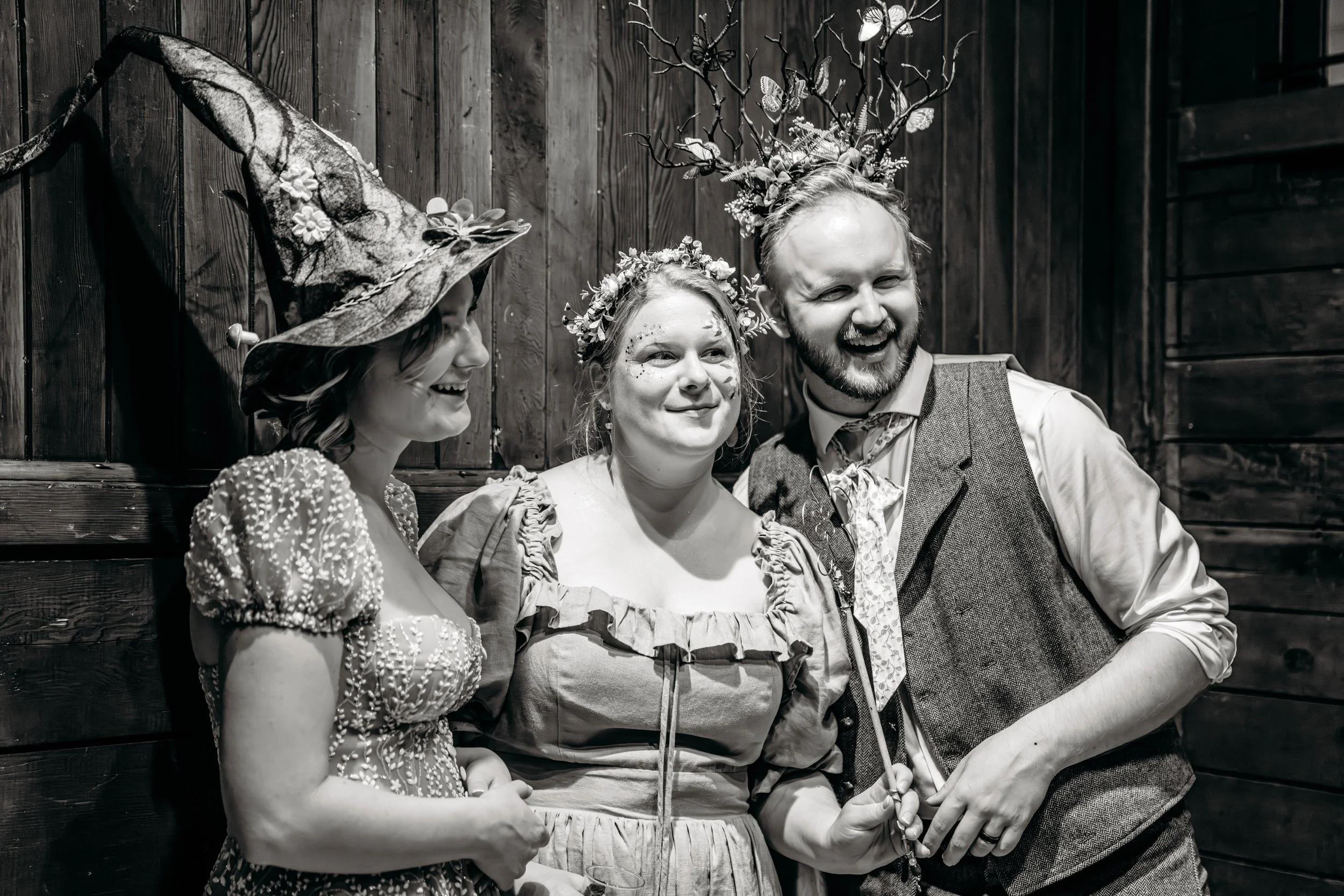 Three people stand closely together, smiling and enjoying each other's company against a wooden wall. The woman on the left wears a whimsical, textured dress and a large, decorated witch hat. The woman in the middle wears a floral crown, striped dres