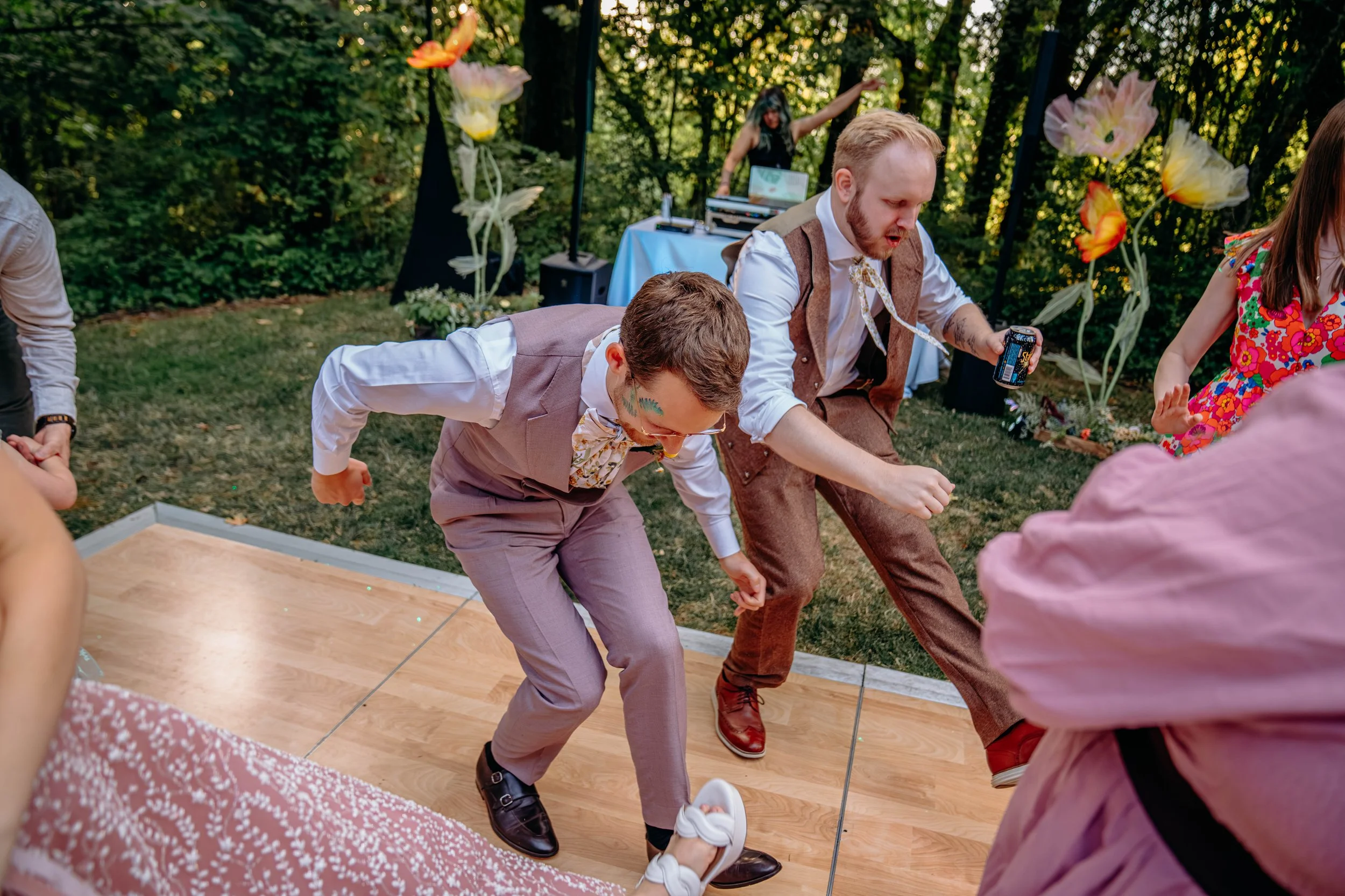 People dancing on a wooden dance floor outdoors at a party, with two men in suits in the foreground and a woman in the background near a DJ.