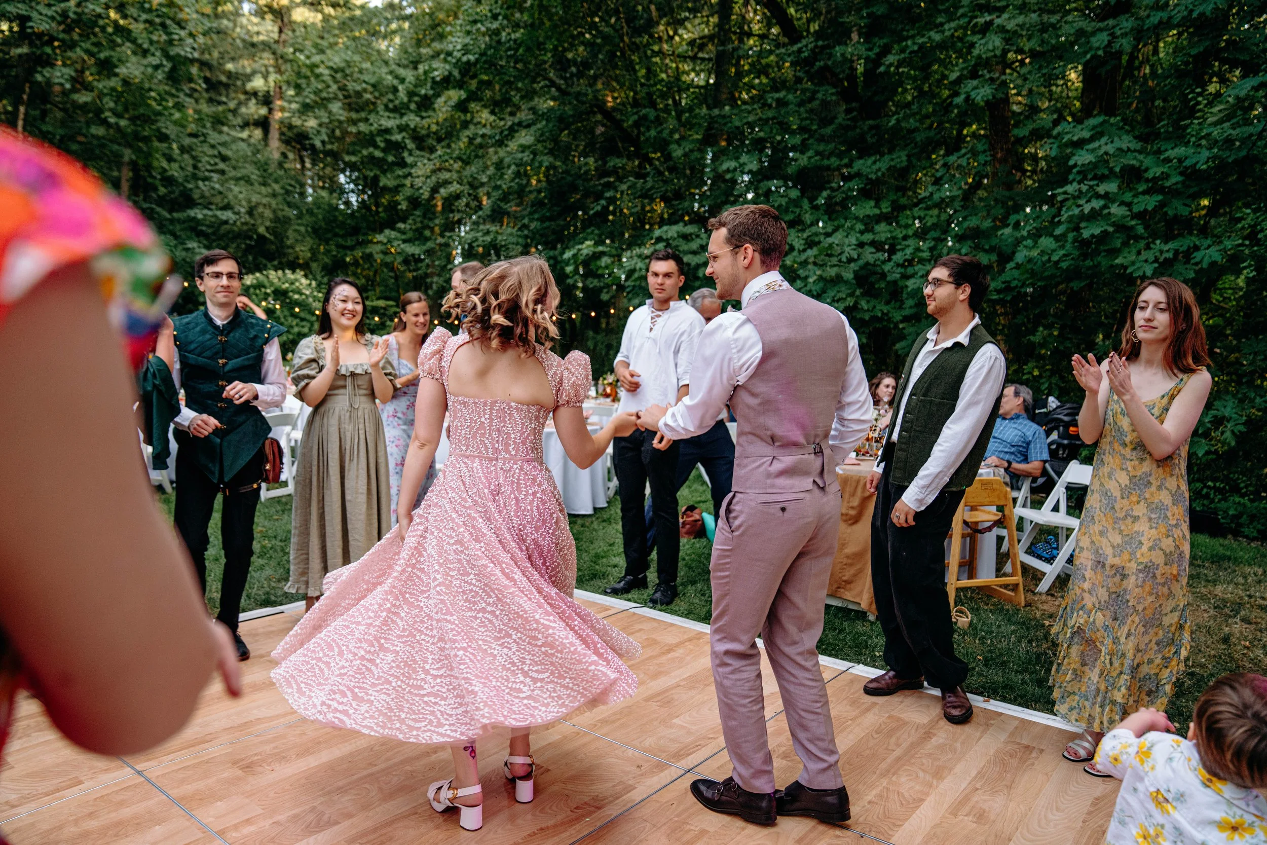 People dancing and socializing at an outdoor event surrounded by trees, with some guests clapping and smiling.