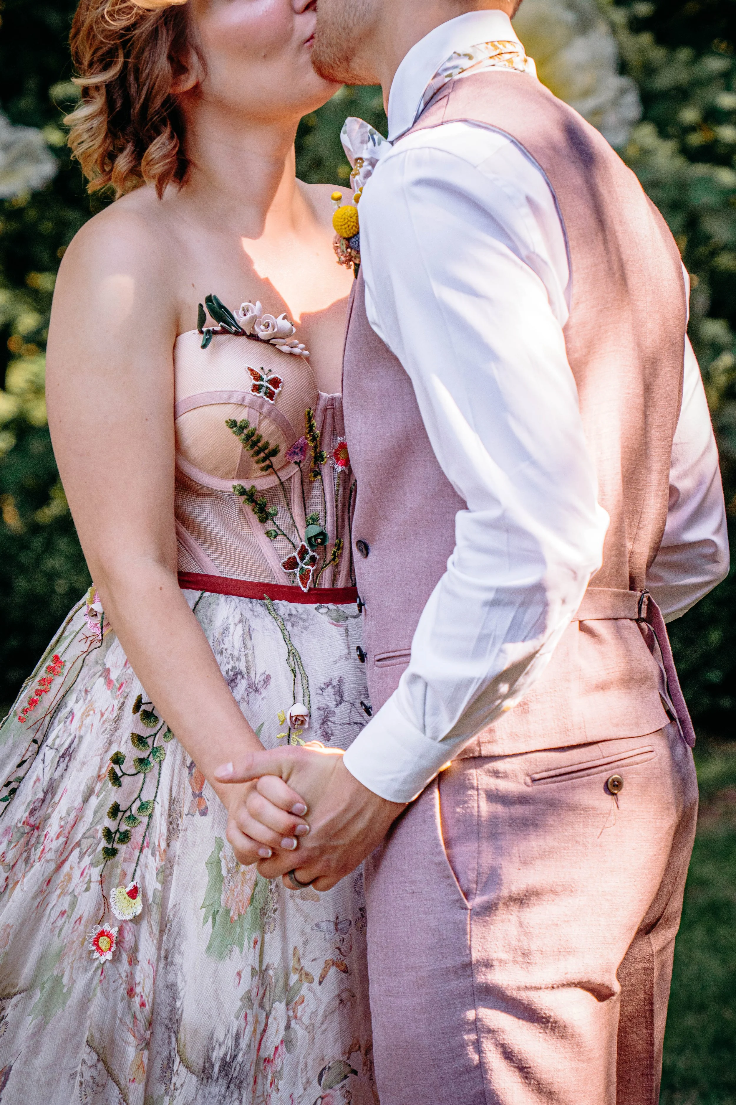 A couple holding hands and kissing outdoors, dressed in wedding attire with floral accents.