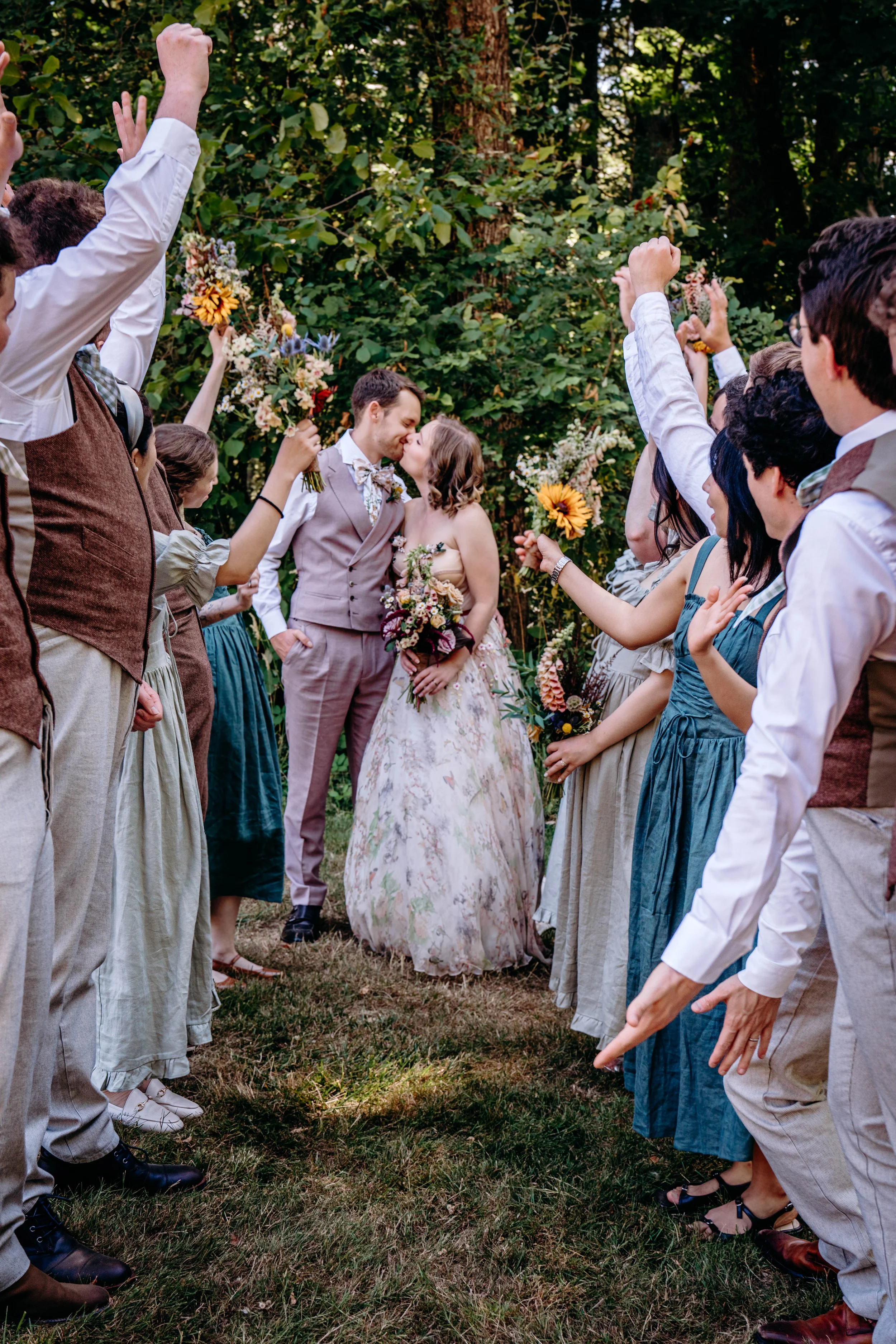 A bride and groom sharing a kiss while surrounded by friends and family holding bouquets, outdoors in a wooded area during their wedding celebration.