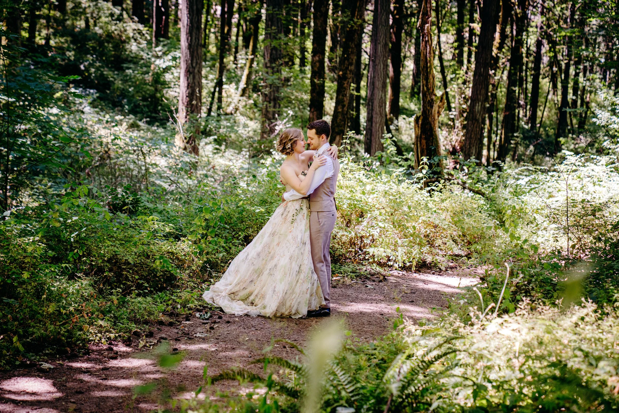 A wedding couple embracing in a forest with tall trees and green foliage.