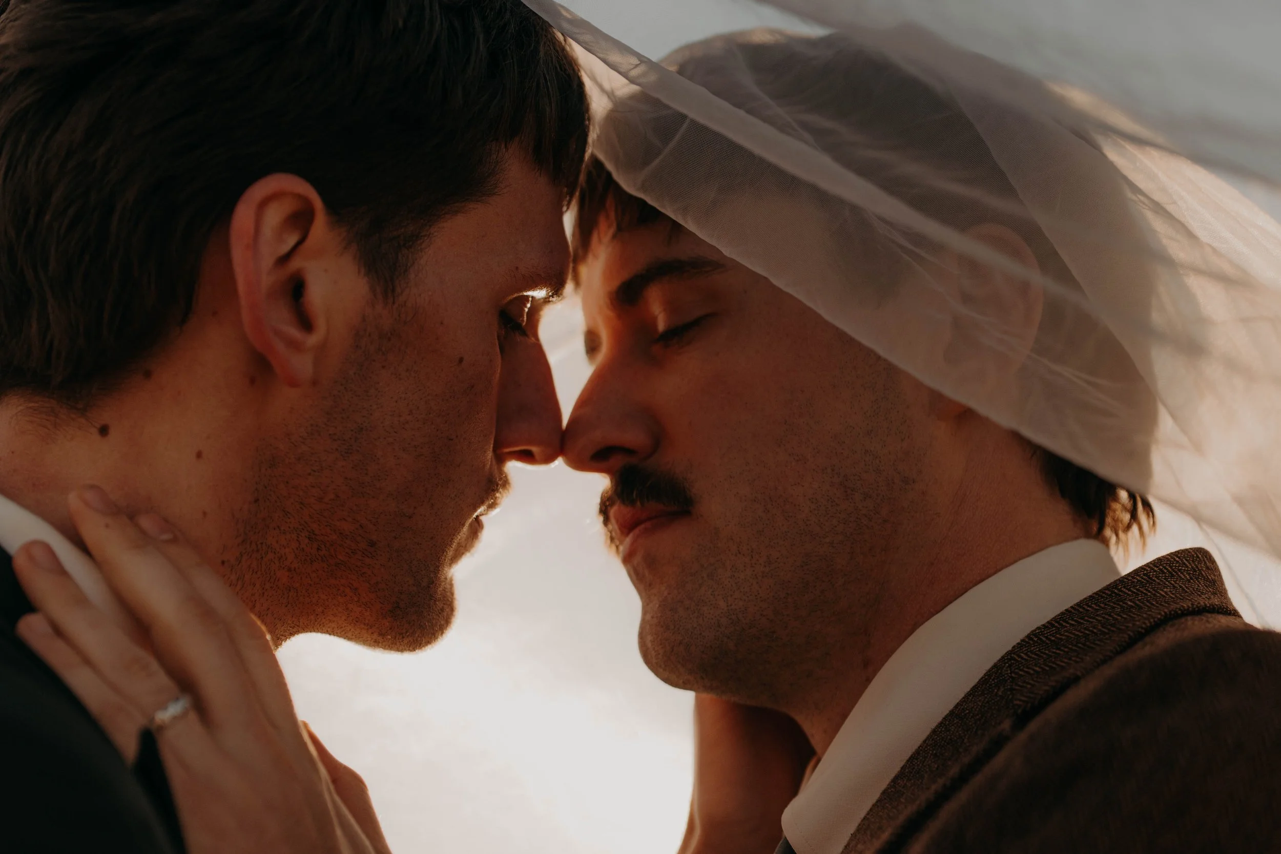 Two men with closed eyes and touching foreheads in a close embrace, wearing wedding attire, with sunlight in the background.