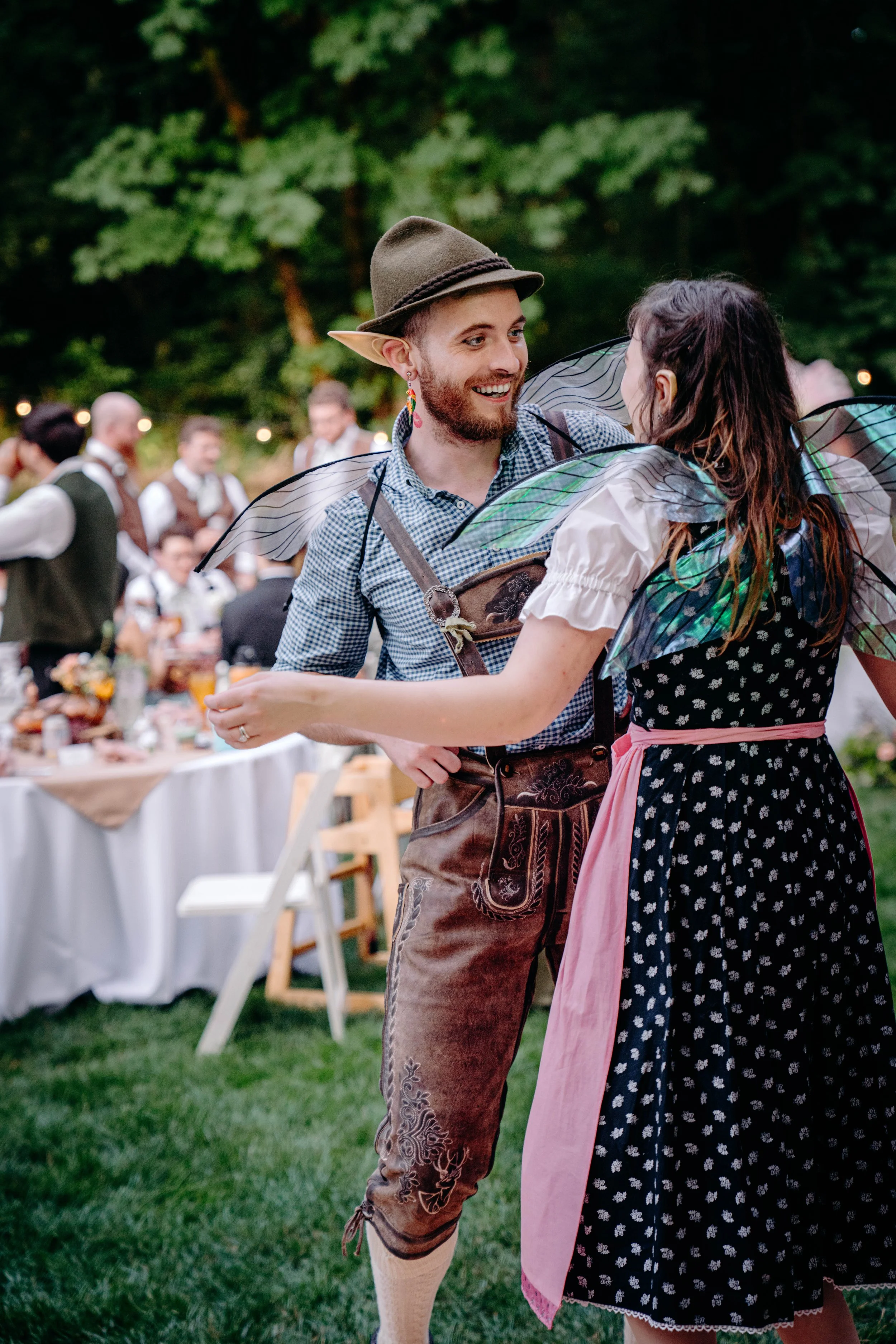 A man and woman dressed in traditional German costumes, dancing and smiling at an outdoor event with fairy lights and guests in the background.