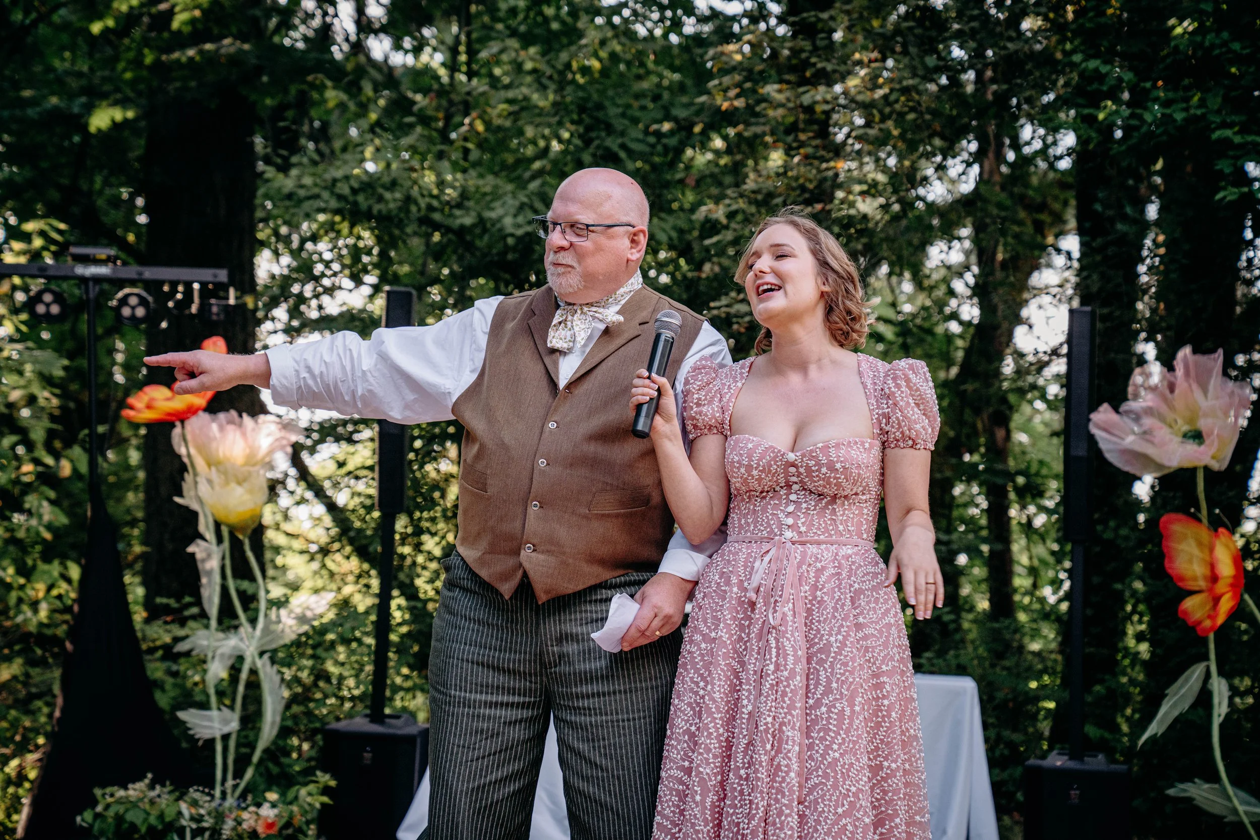 A man and a woman standing outdoors, smiling and engaging with an audience. The man is pointing to the left, holding a tissue in his left hand, and dressed in a brown vest and striped trousers. The woman is holding a microphone, wearing a pink patter