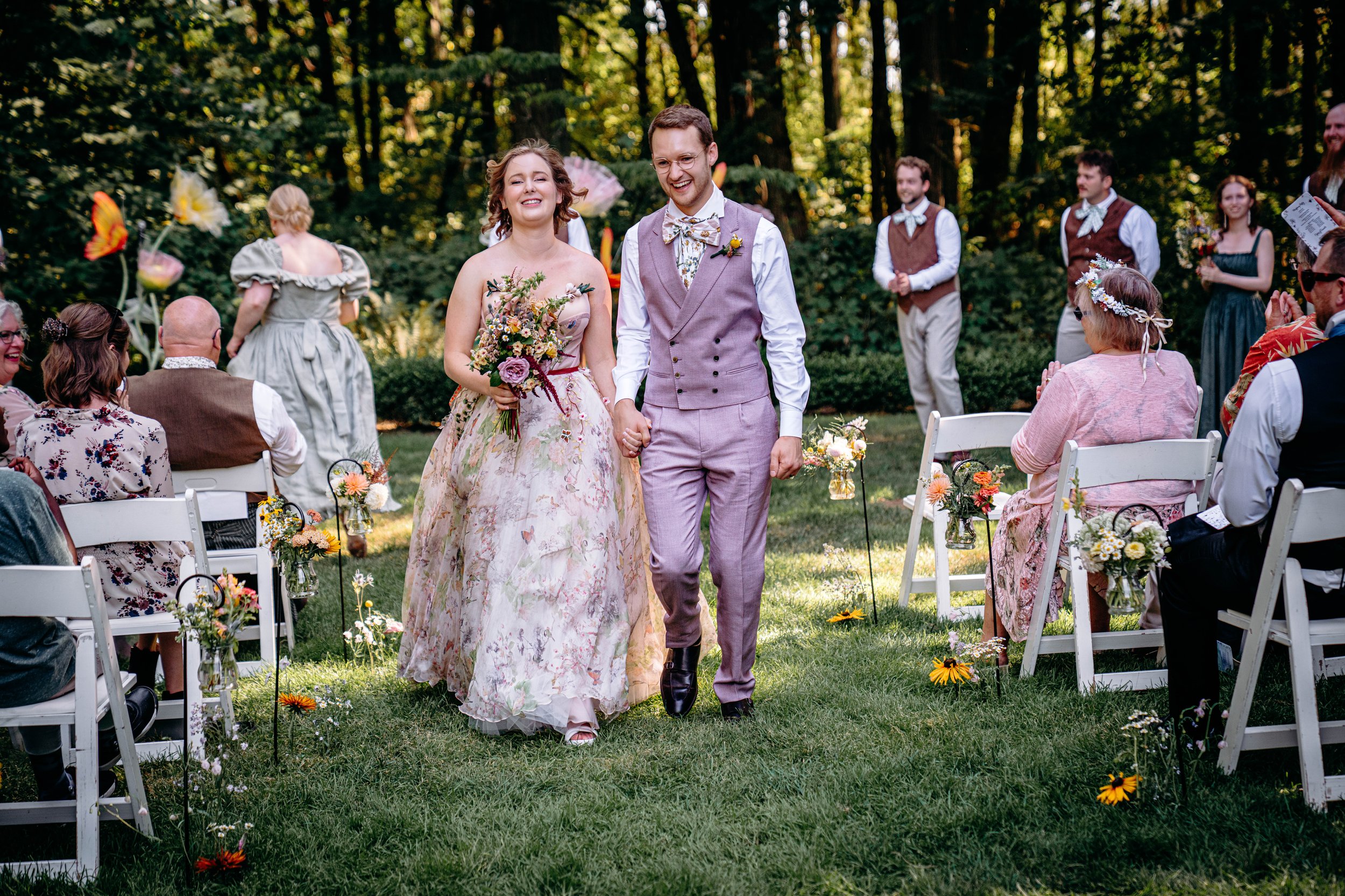 A newly married couple walking down the aisle at an outdoor wedding ceremony, holding hands, with guests seated on either side, surrounded by trees and flowers.