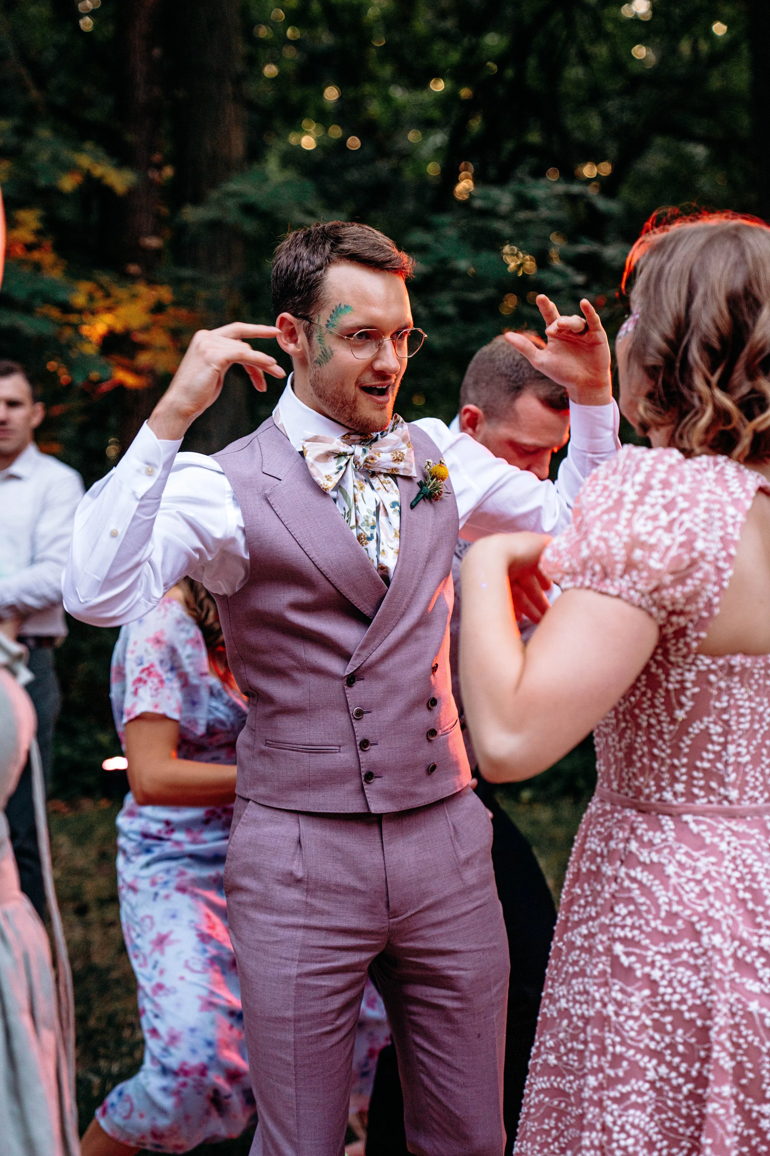 Man dancing at an outdoor party, wearing a light purple suit, patterned bow tie, glasses, and face paint of green leaves, with other guests in casual attire around. Forest backdrop with warm lighting.