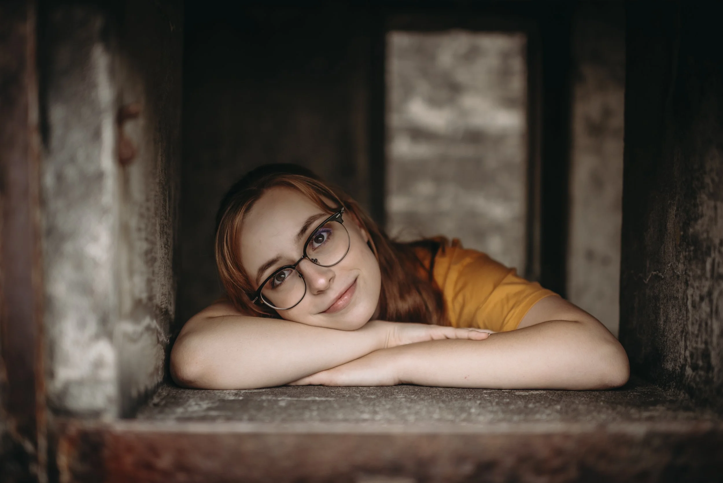A young woman with red hair and glasses resting her head on her crossed arms, lying inside a dark, narrow brick or stone enclosure, looking at the camera with a gentle smile.