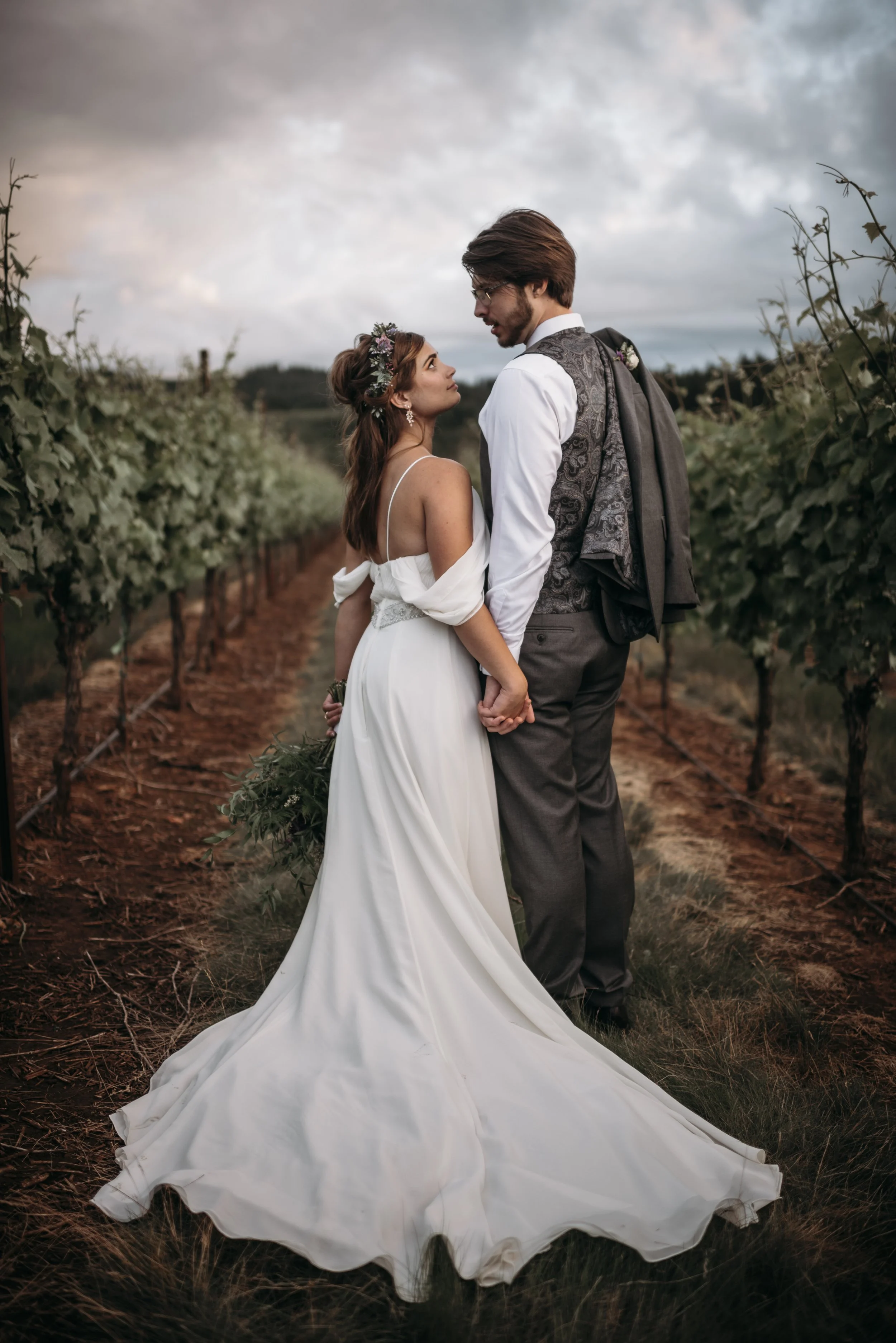 A bride and groom standing in a vineyard, holding hands and looking into each other's eyes, with cloudy sky overhead.