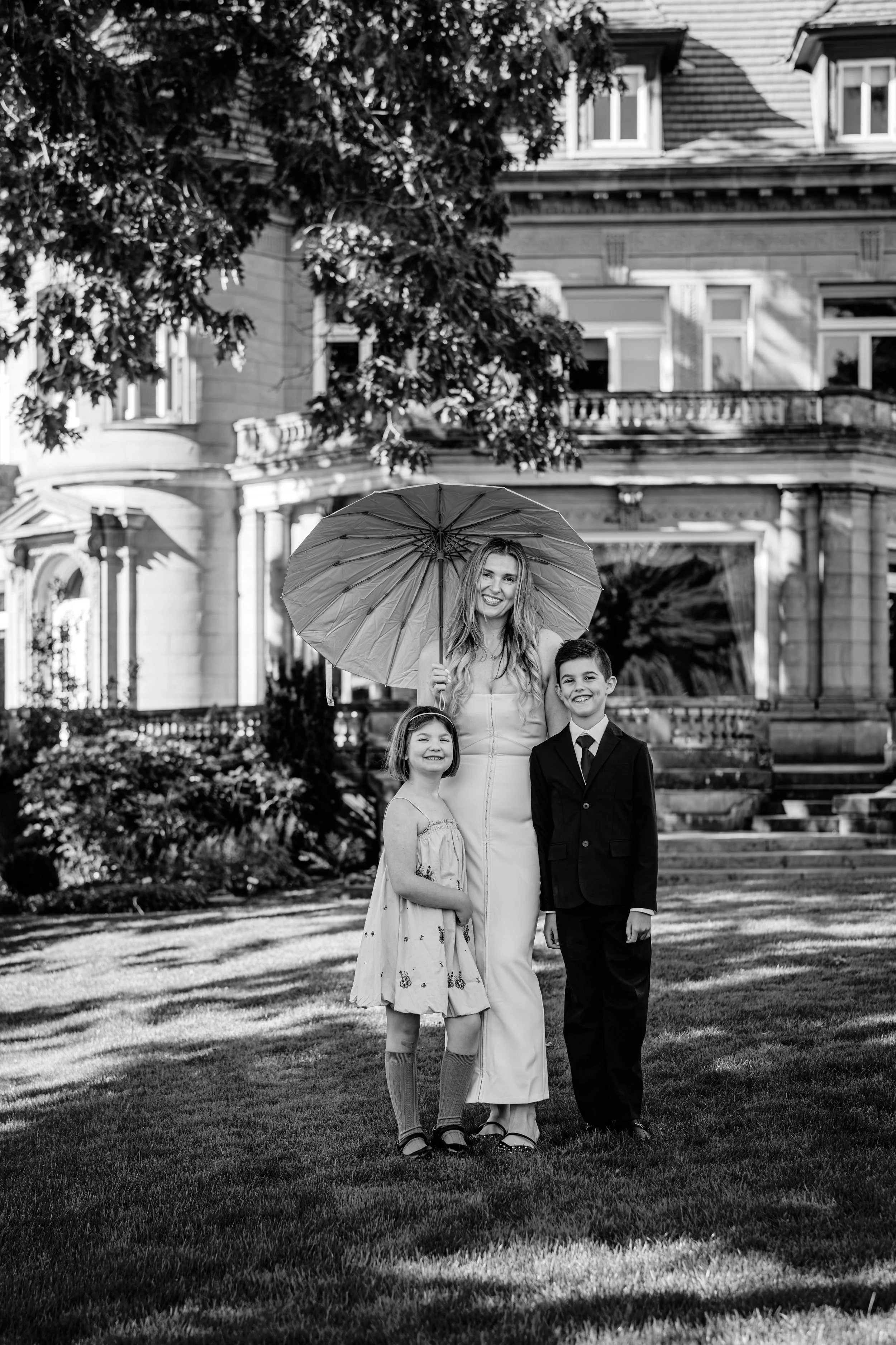 A woman and two children standing on a lawn, smiling at the camera. The woman is holding an umbrella, and they are in front of a large house with ornate architecture.