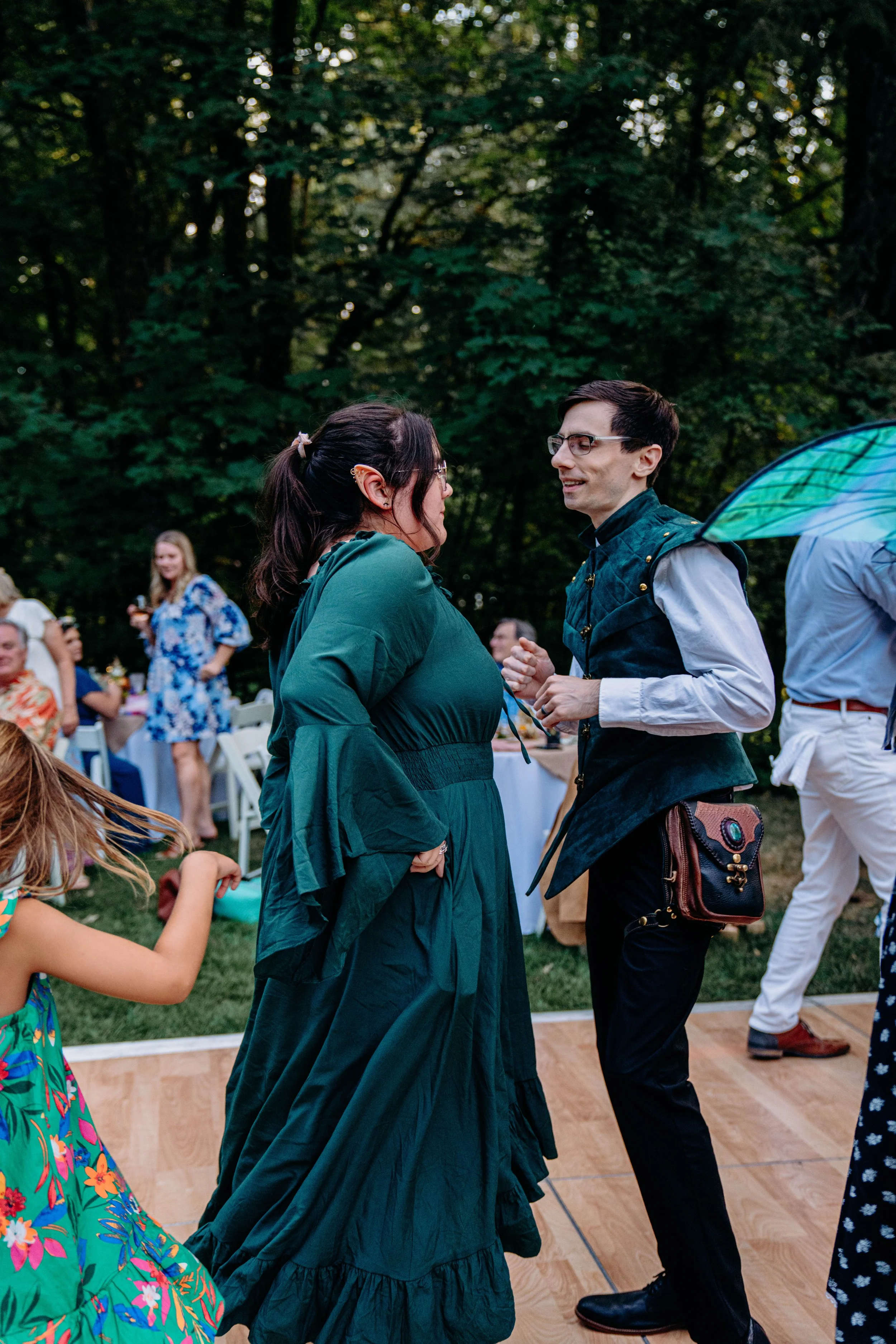 A group of people dancing and socializing outdoors at a party or celebration in a wooded area with tables and chairs in the background.