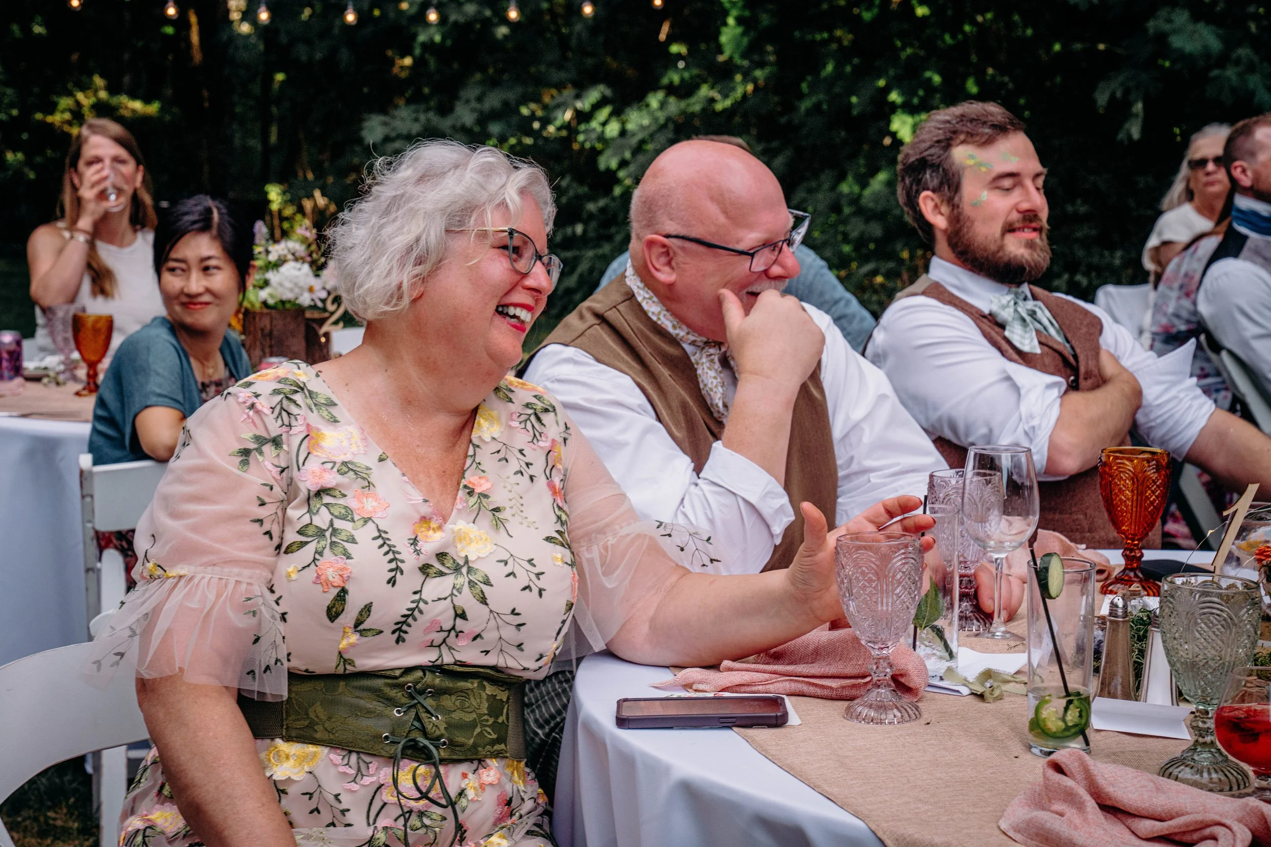 People enjoying a wedding reception outdoors, sitting at a decorated table, laughing and smiling.