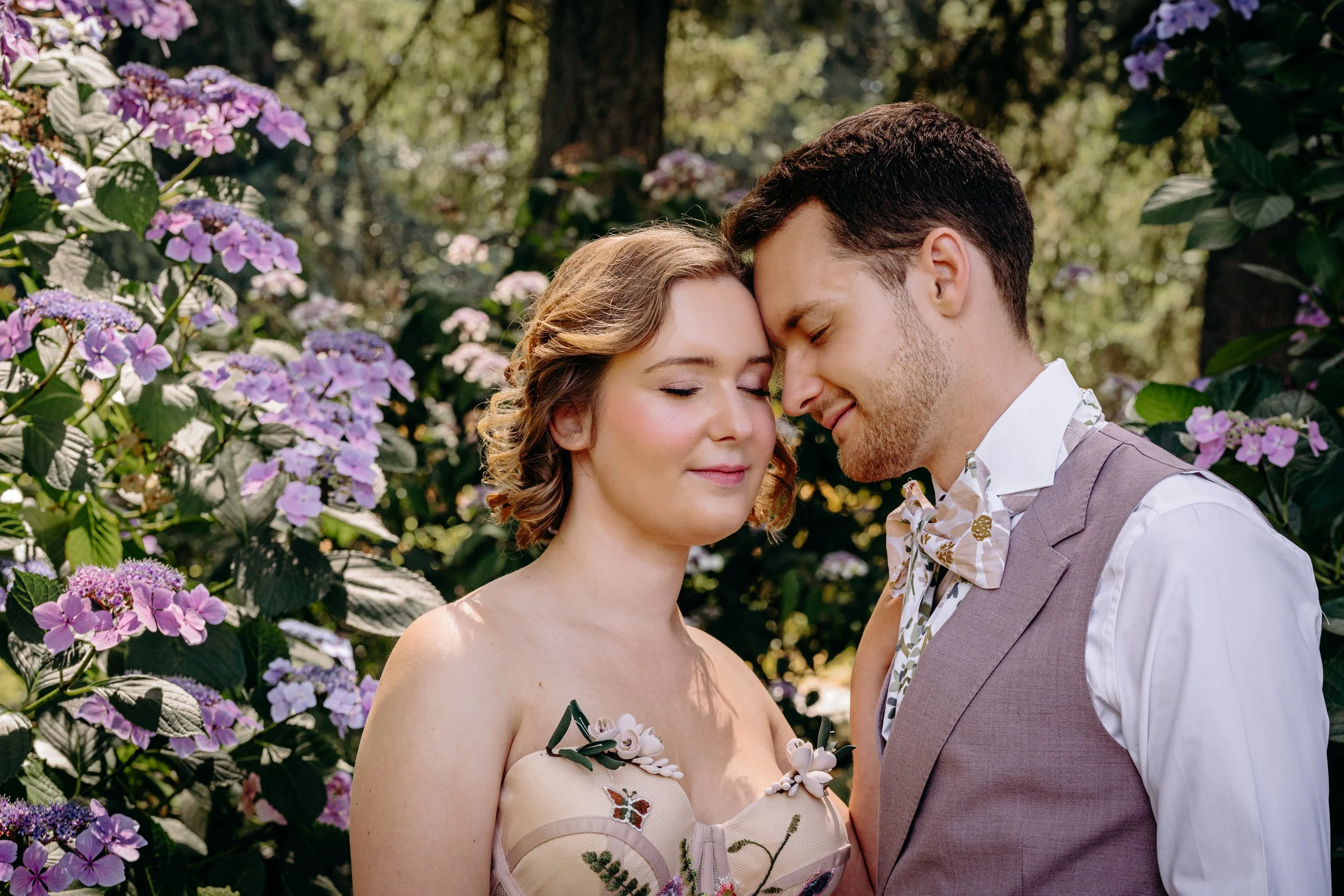 A man and woman in formal attire with foreheads touching, surrounded by purple and pink flowers in a garden.