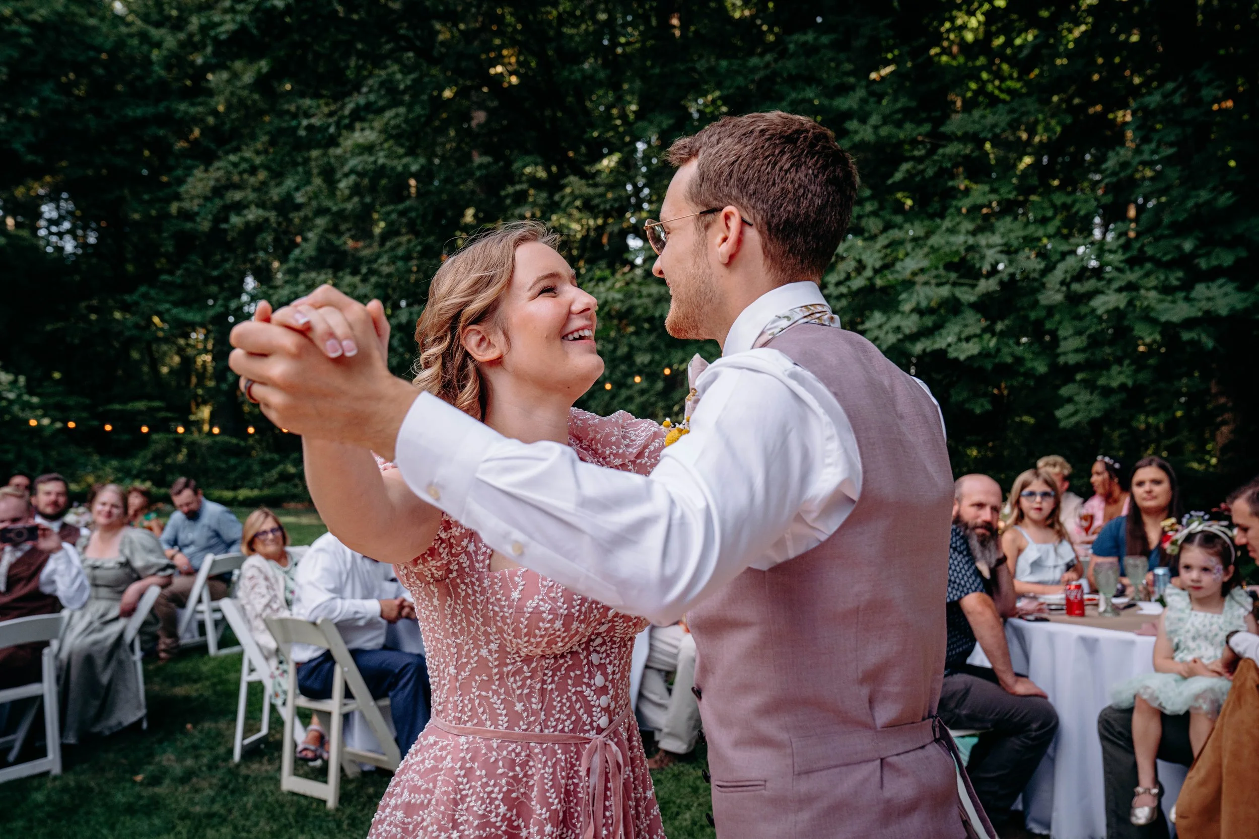 Couple dancing at an outdoor wedding reception, smiling and looking into each other's eyes, with guests seated at tables in the background.