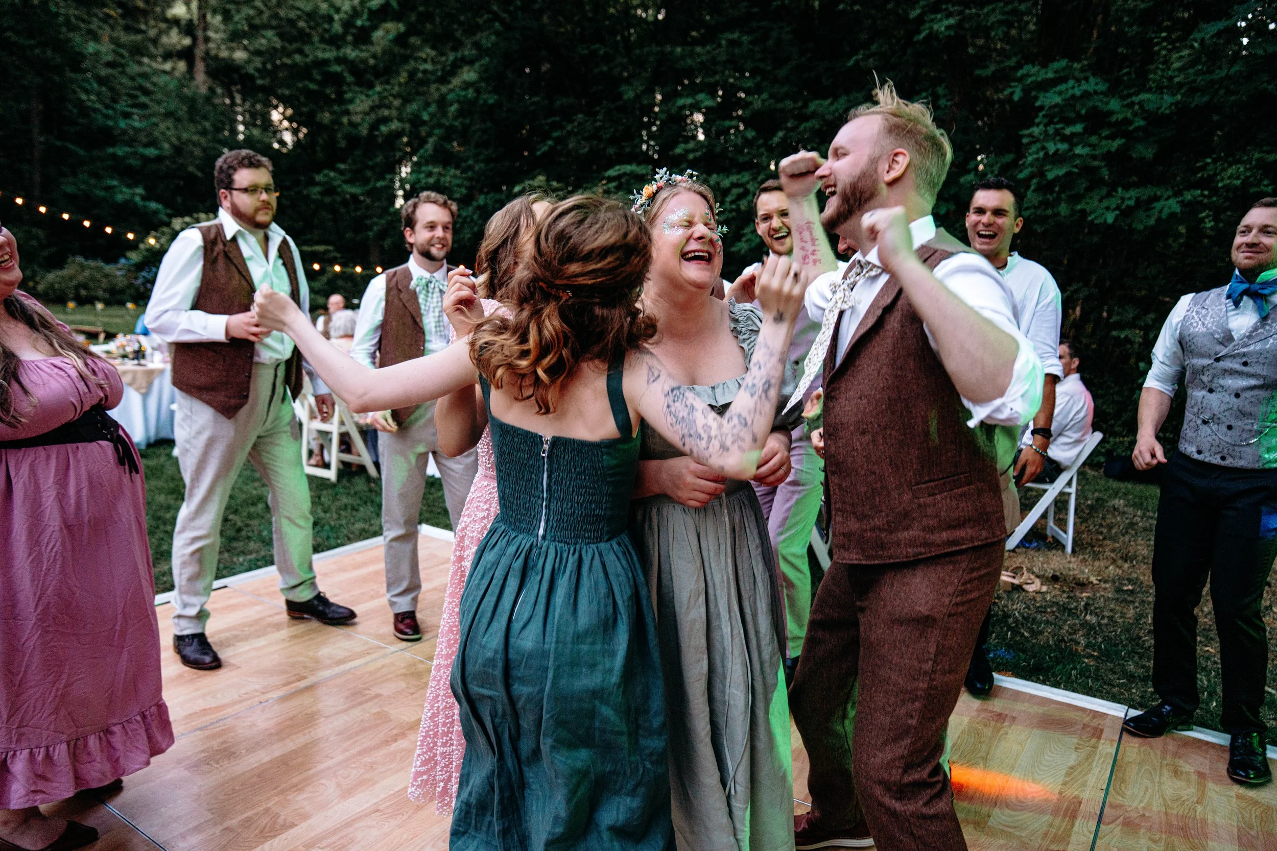 People dancing and celebrating outdoors at a wedding reception, with smiles and laughter, in a wooded area during the evening.