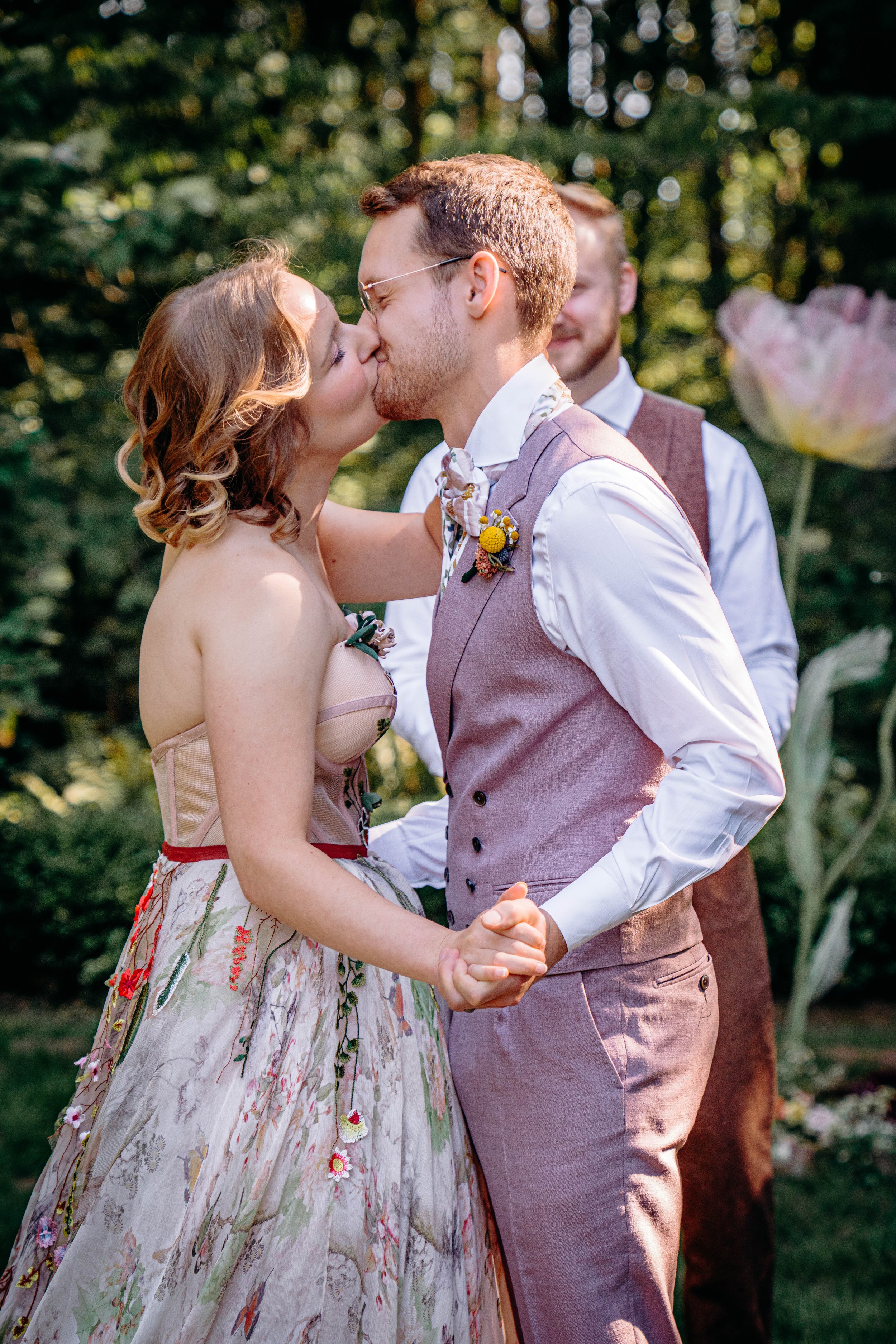 A couple kissing during their wedding ceremony outdoors, with a man in the background and lush greenery around them.