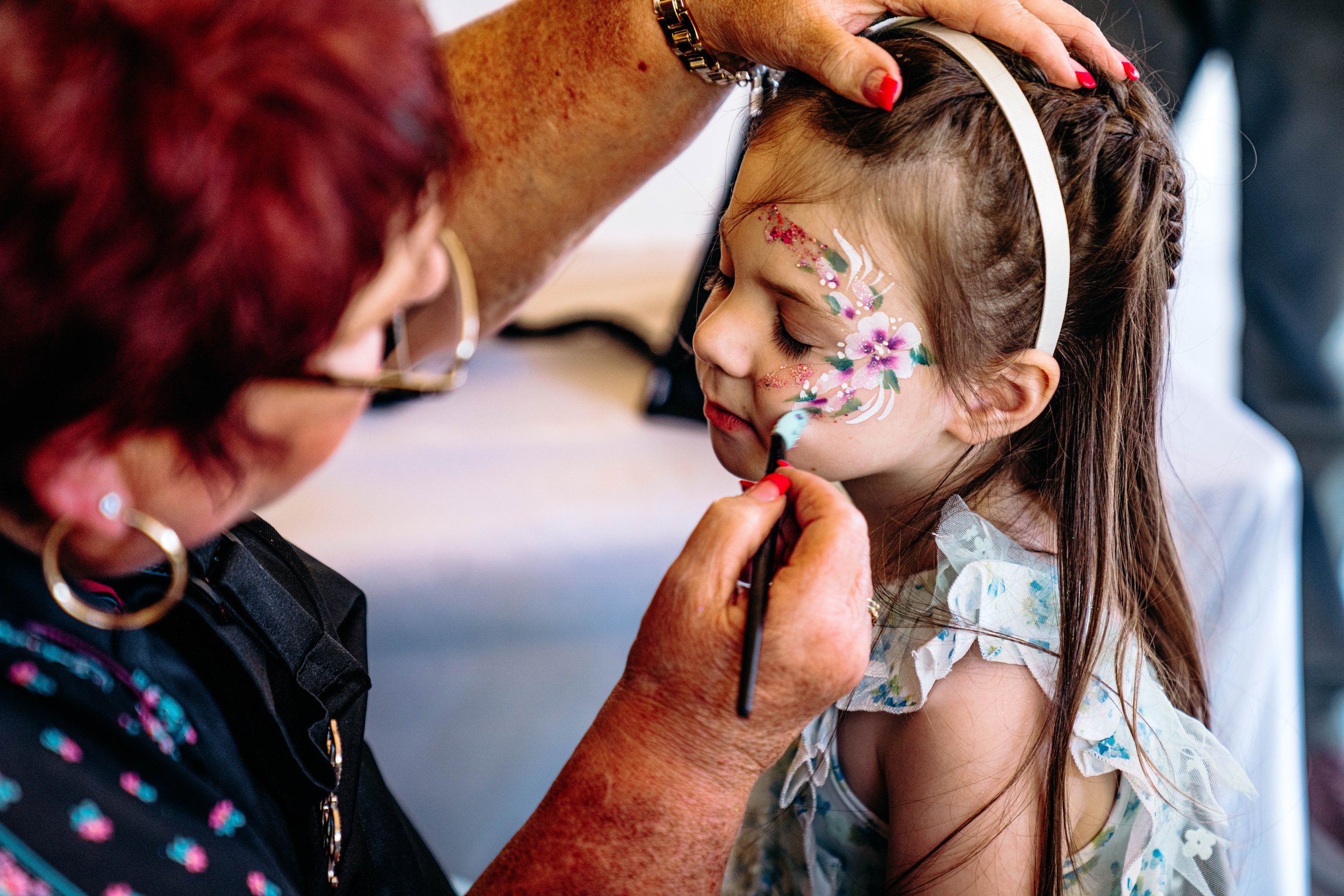 An elderly woman is painting a floral face design on a young girl's face during a face painting activity.