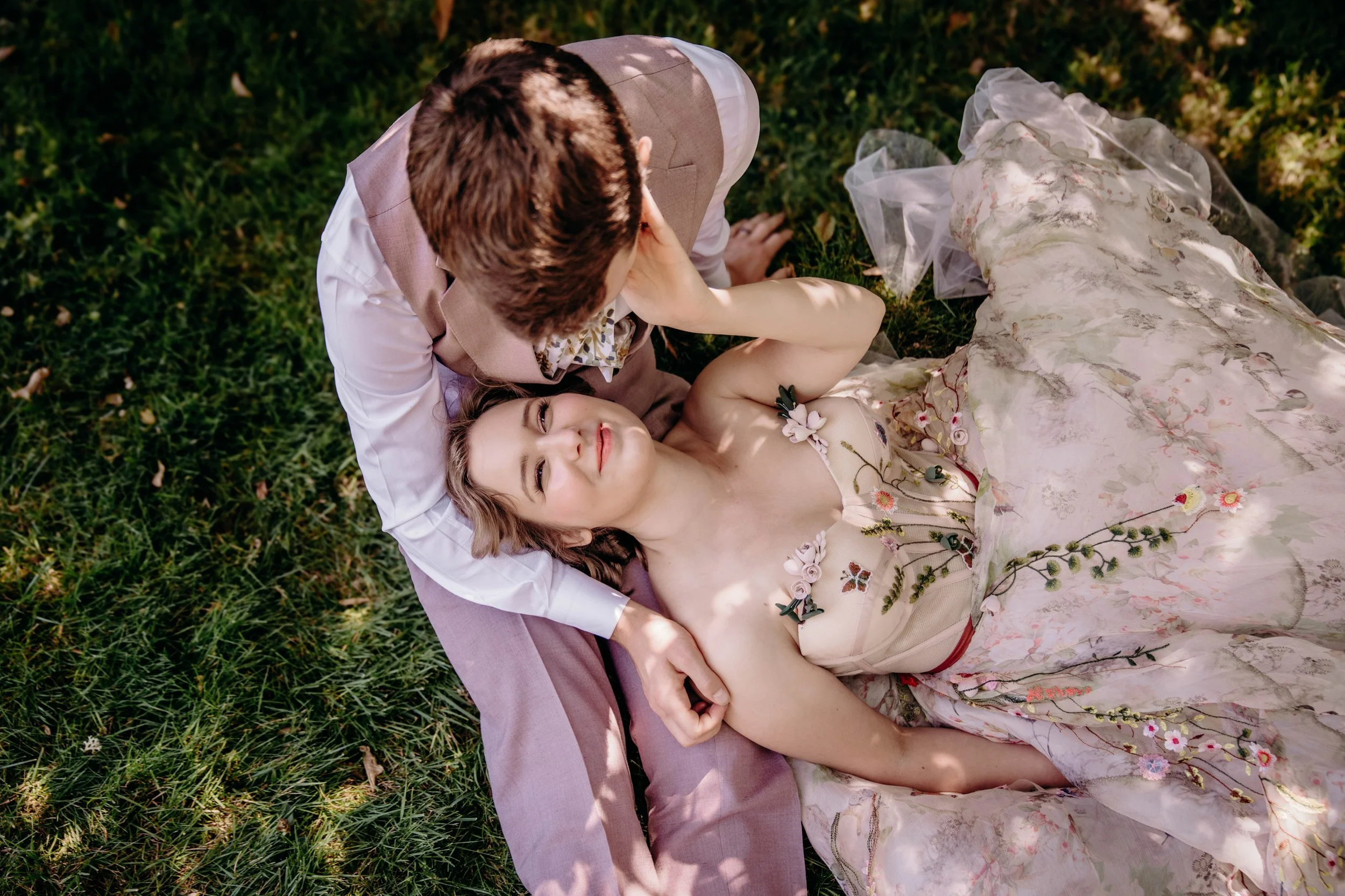 A woman lying on her back on a grassy field, wearing a wedding dress with floral embroidery, and a man kneeling beside her, leaning over and gently touching her face.