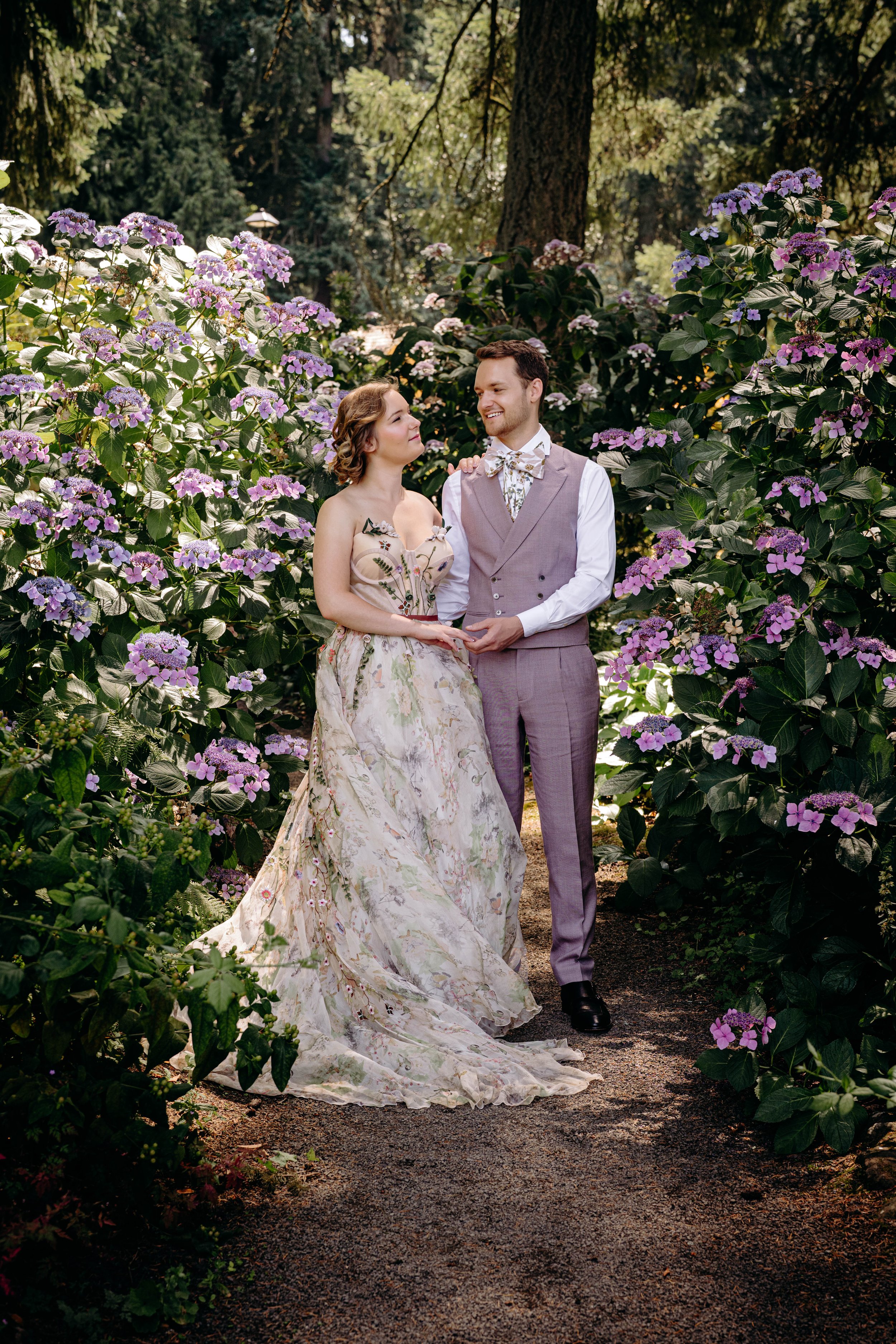 A couple dressed in vintage-inspired wedding attire standing among flowering purple and pink plants in a wooded outdoor setting, gazing at each other.