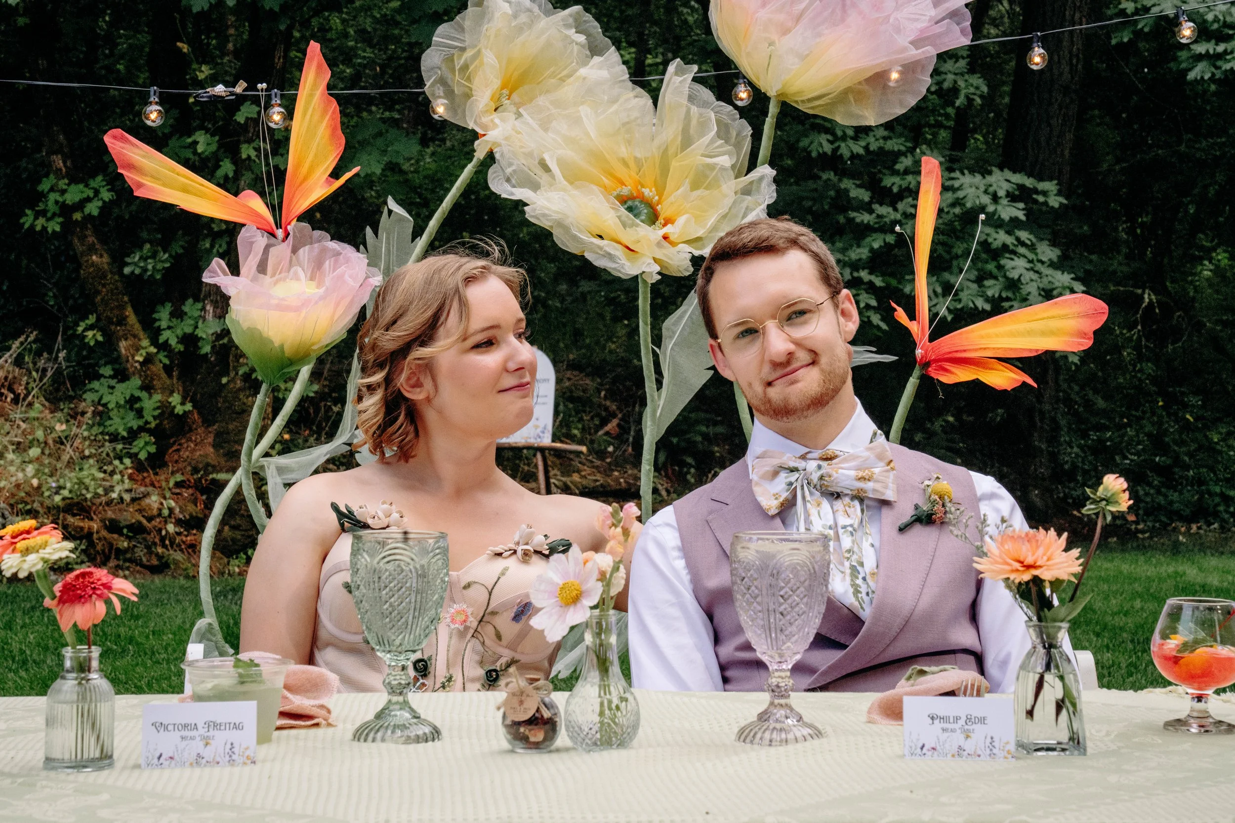A man and woman sit at a decorated table with large artificial flowers and butterfly decorations hanging in the background during outdoor event.