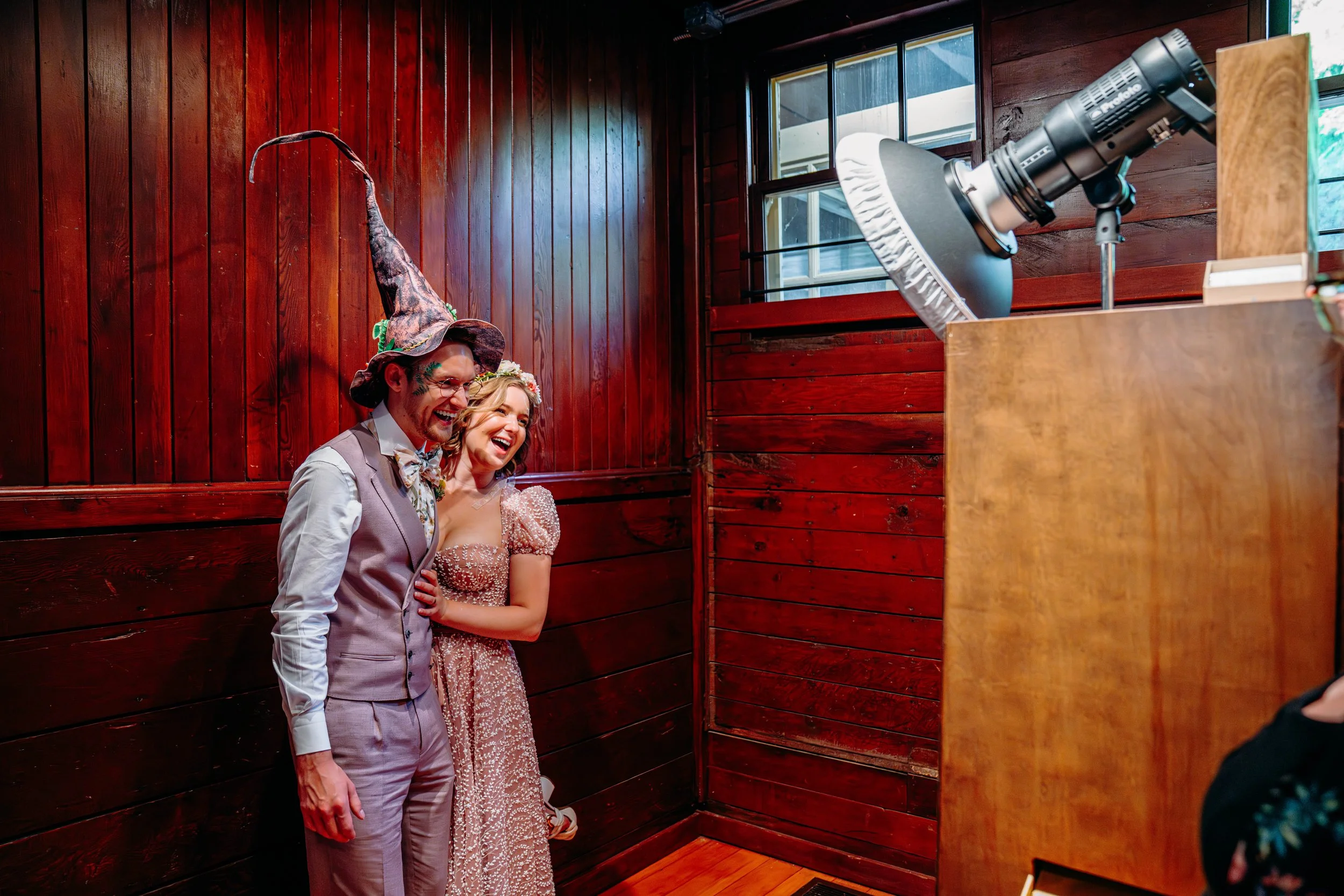 A man in a suit and a woman in a dress are laughing and hugging while taking photos in a photo booth, with a large camera setup in front of them.