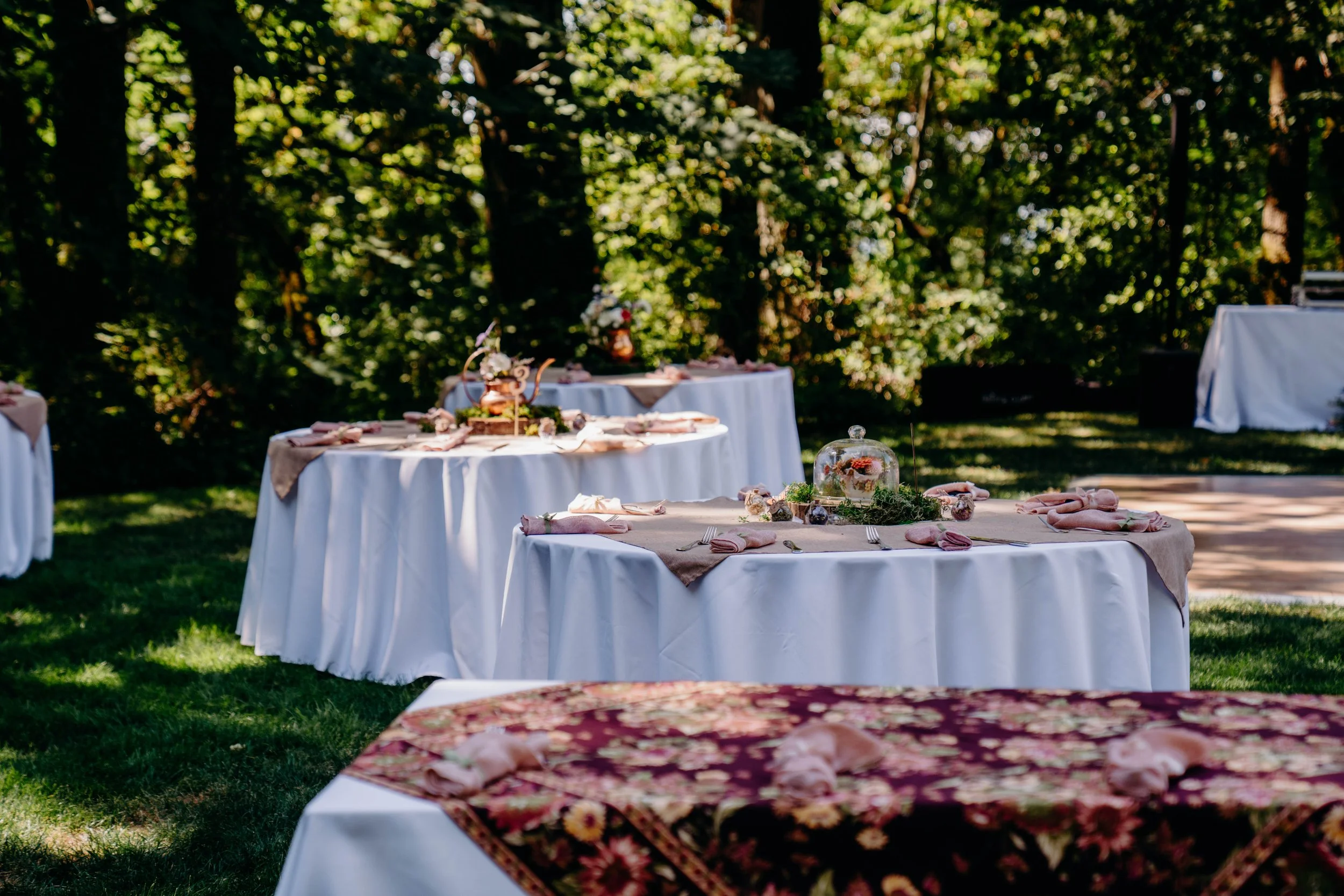 Outdoor event setup with round tables covered in white tablecloths and decorated with pink napkins, flower arrangements under glass domes, and a wooded background with sunlight filtering through trees.