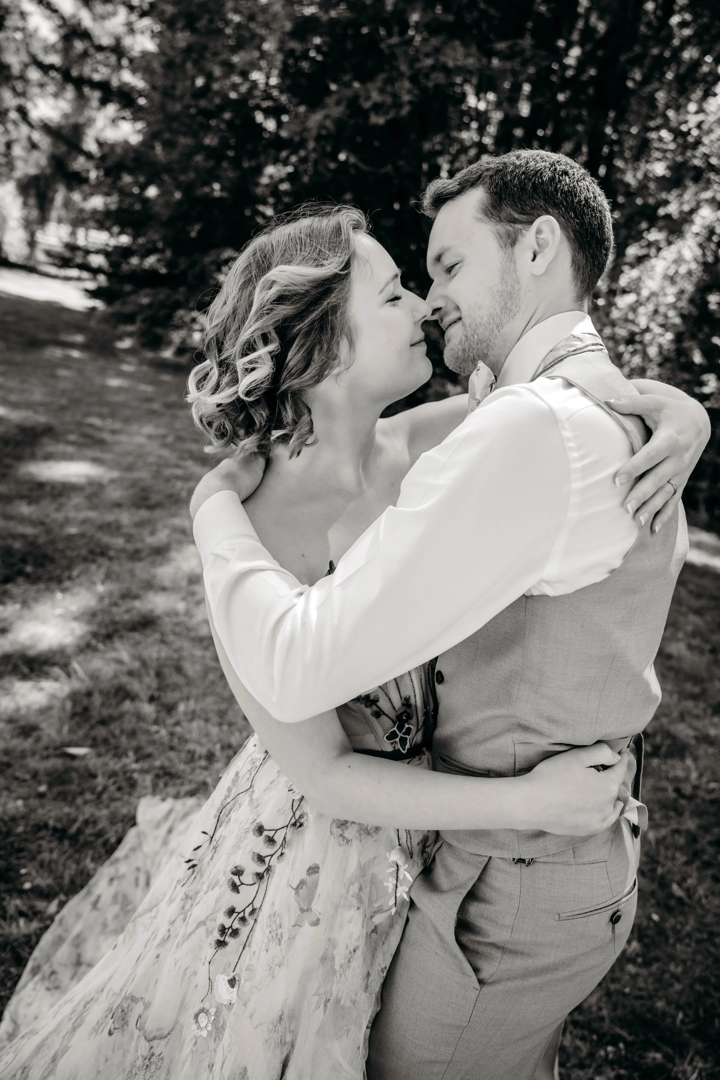 A black-and-white photo of a couple embracing outdoors, their faces close together, with trees in the background.