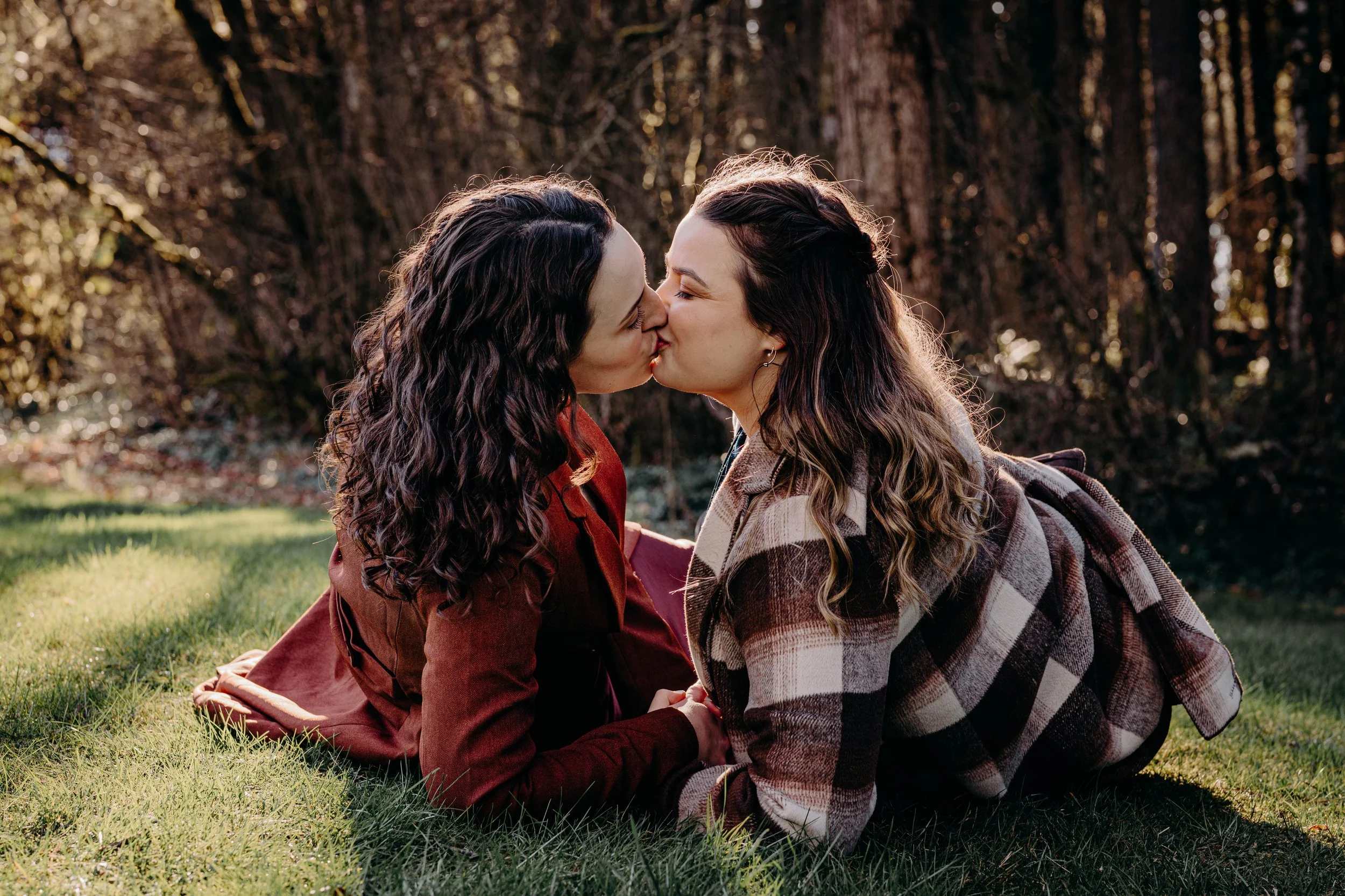 Two women with curly hair kissing on the lips while lying on their stomachs on grass in a wooded area.