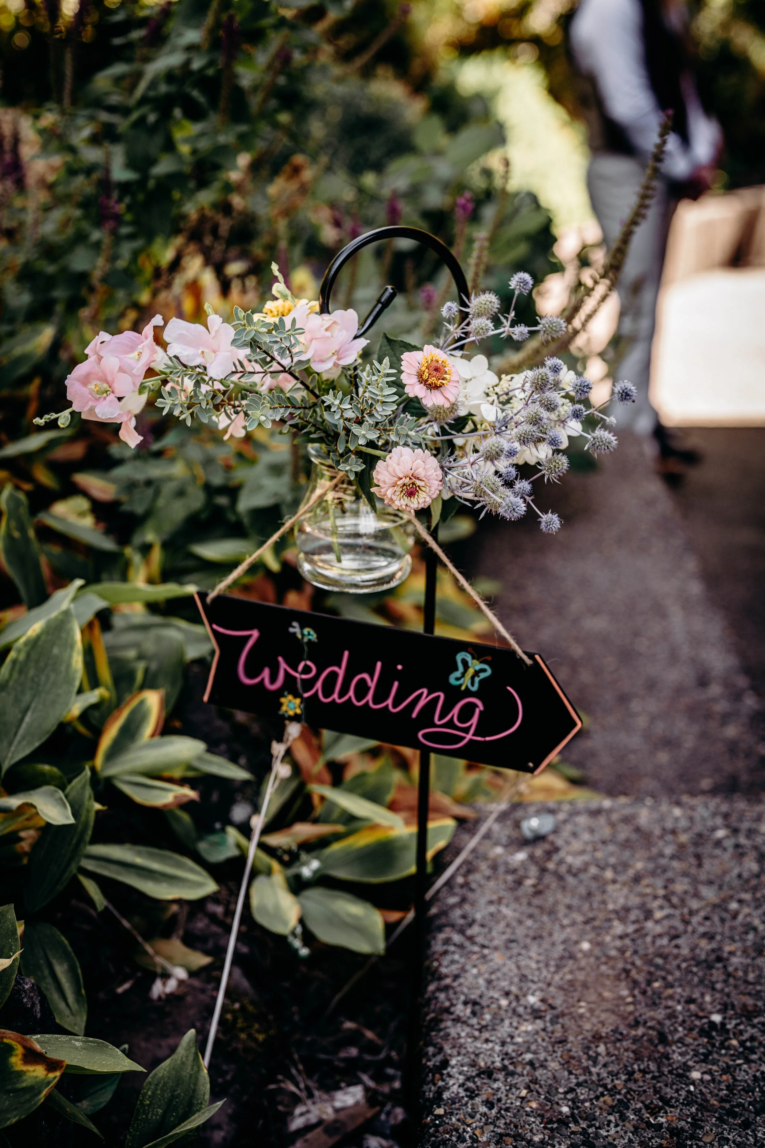 A floral arrangement with pink, purple, and green flowers in a glass jar, placed on a stand next to a black sign with the word 'Wedding' written in pink, hanging on a wire. The arrangement is beside a pathway with greenery in the background.